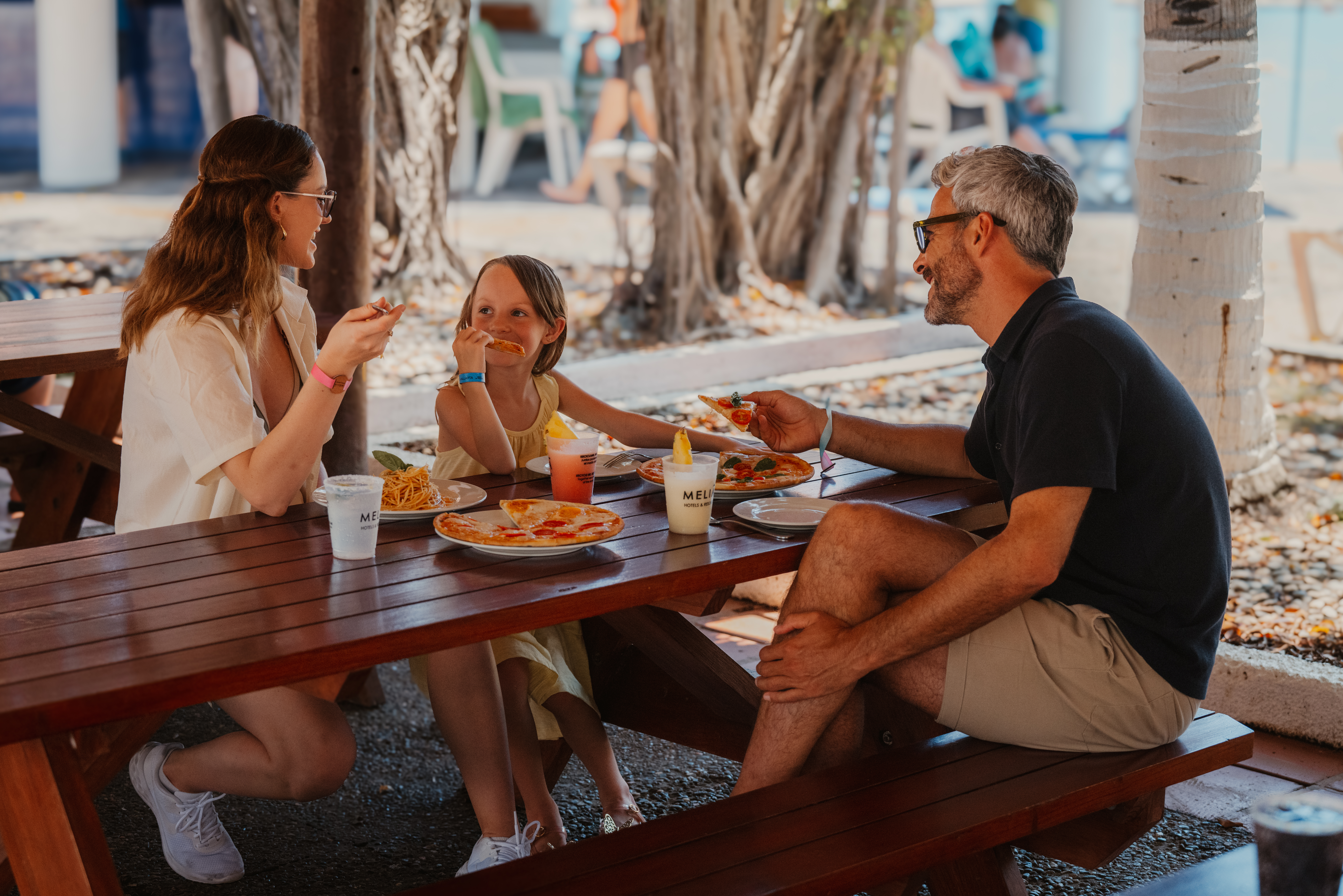 Familia sonriendo comiendo pizza y pasta en resort al aire libre.