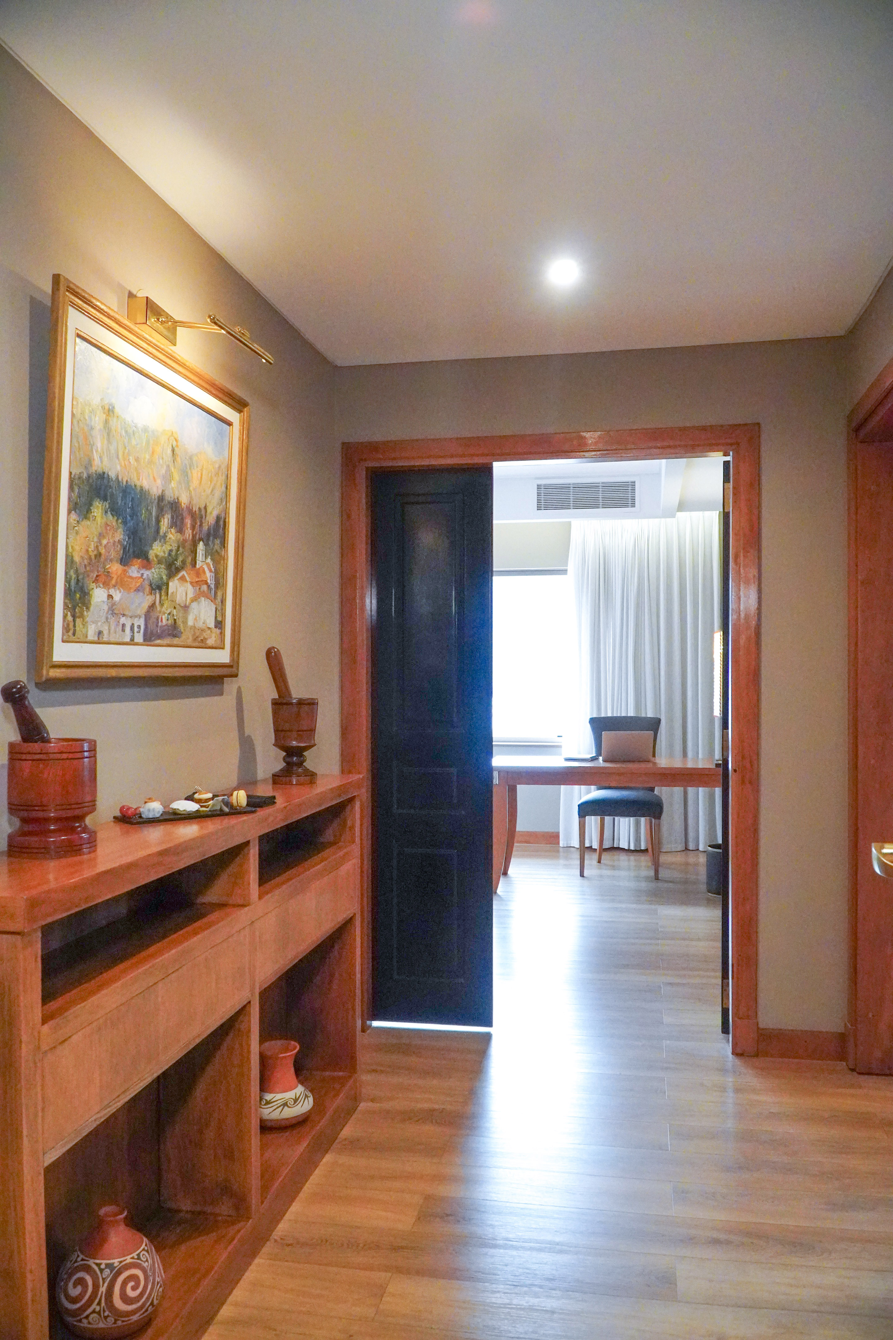 Hotel suite hallway with wooden console, landscape painting, and bright room.