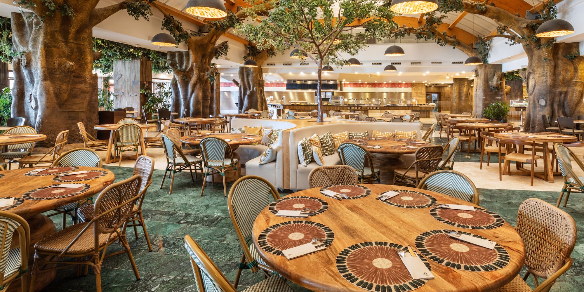 Restaurant interior with large tree columns, wooden tables, and woven chairs.