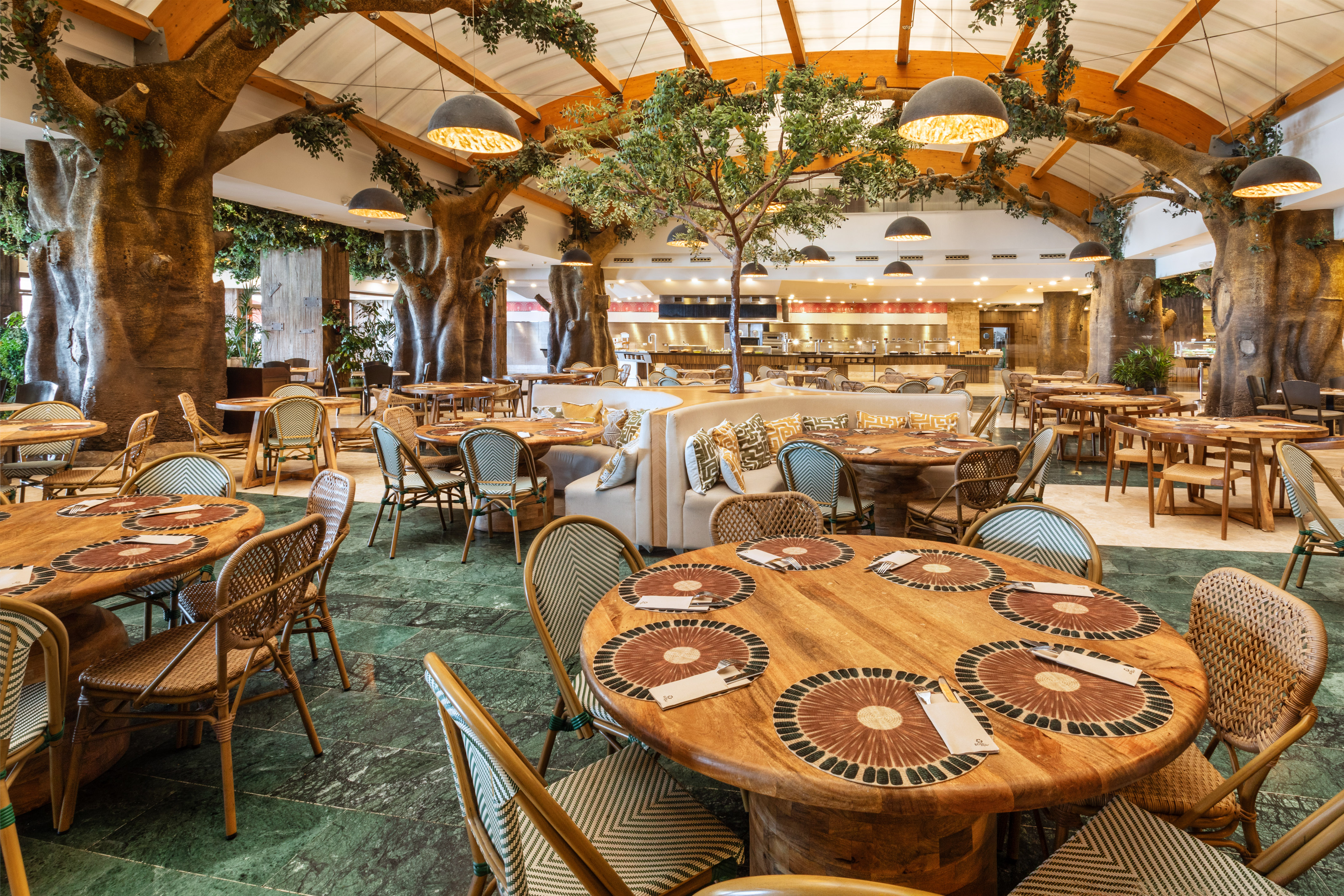 Restaurant interior with large tree columns, wooden tables, and woven chairs.