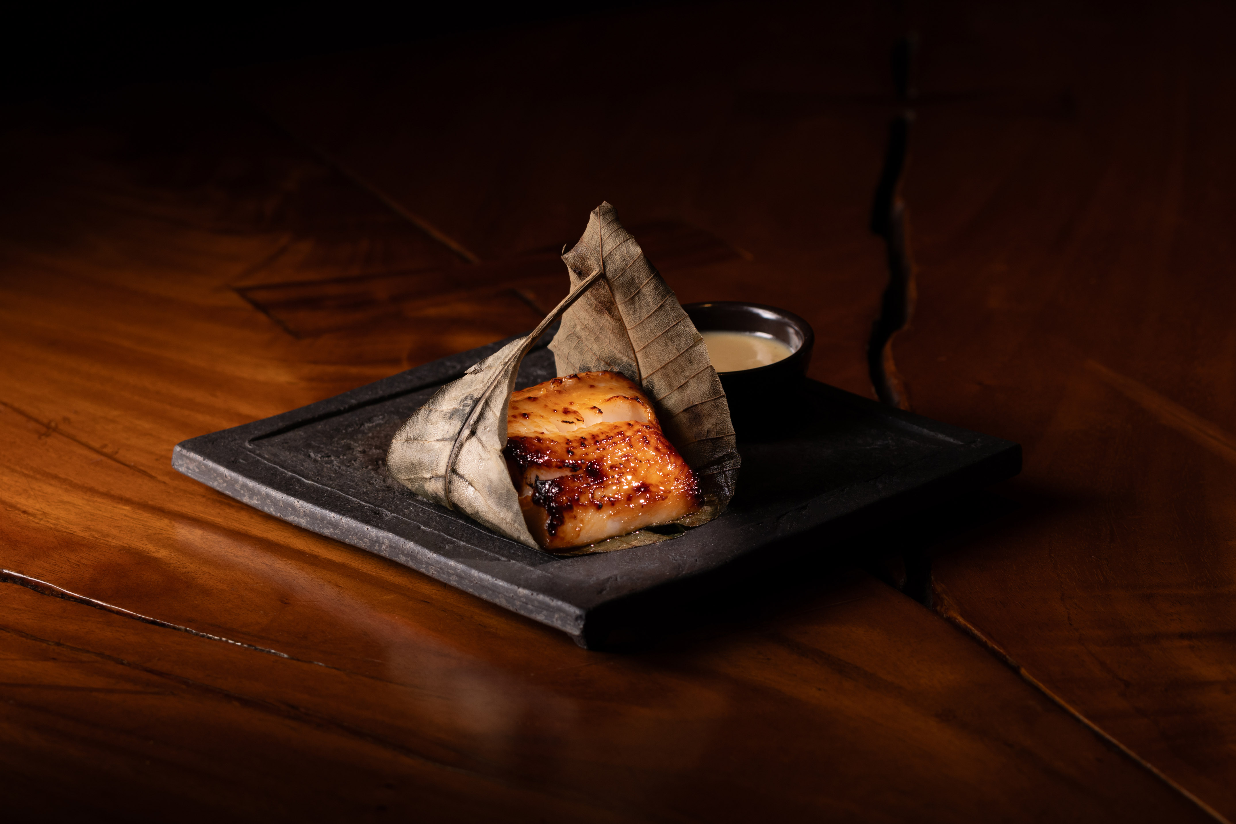 Caramelized miso-glazed fish in a dried leaf on a slate plate with sauce.