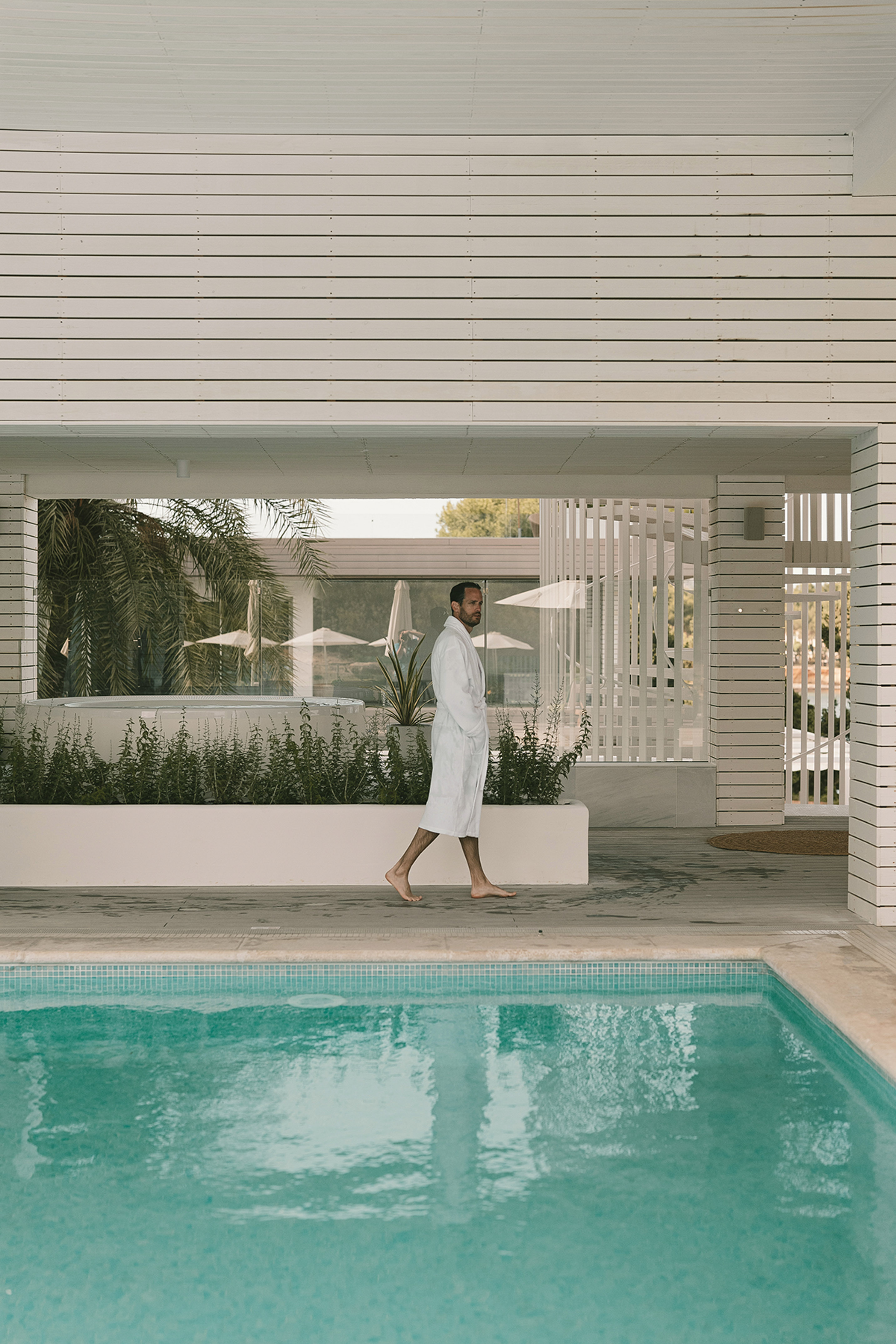 Man in white bathrobe walks barefoot by turquoise resort pool.