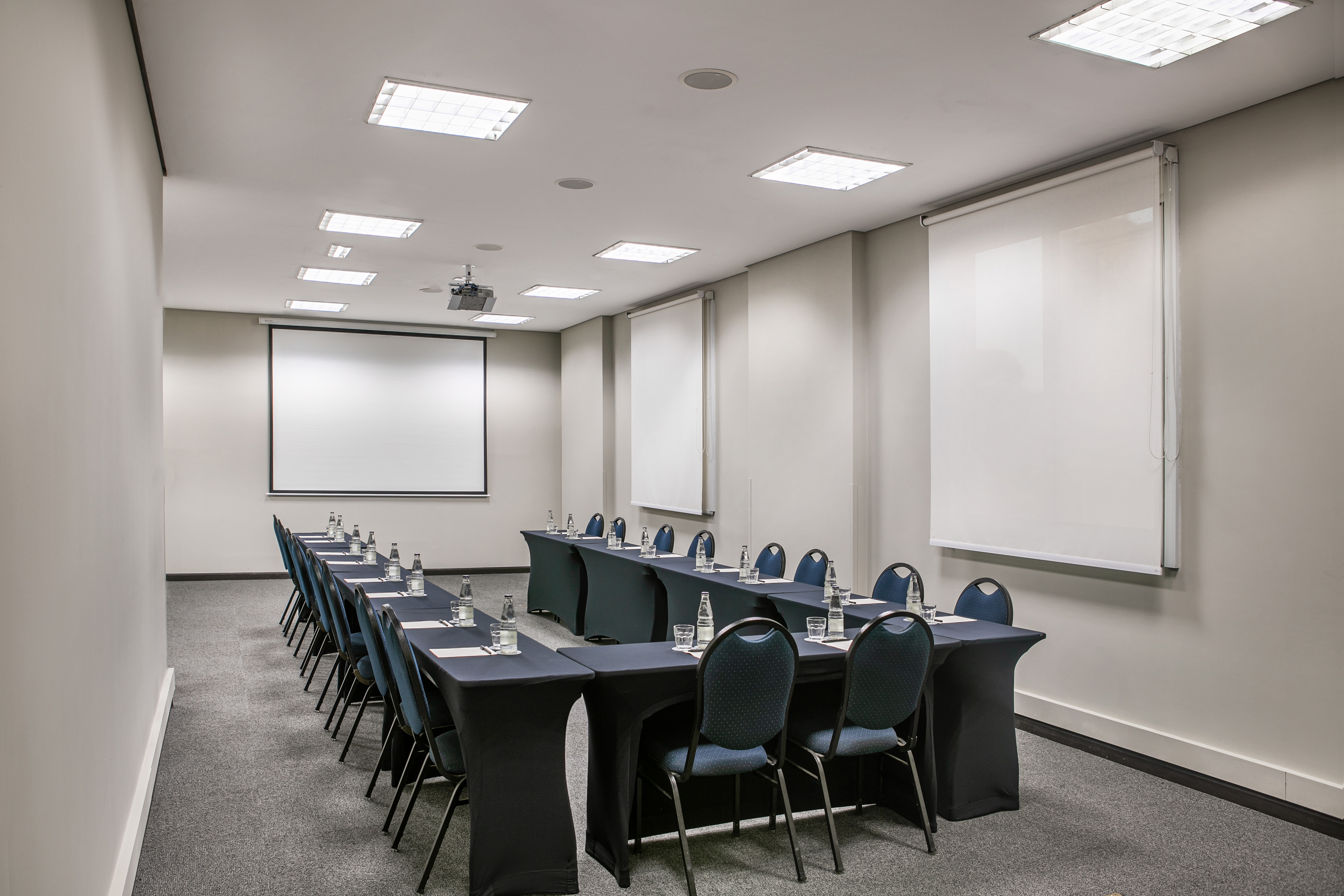 Modern meeting room with U-shaped tables, blue chairs, projector, and screen.