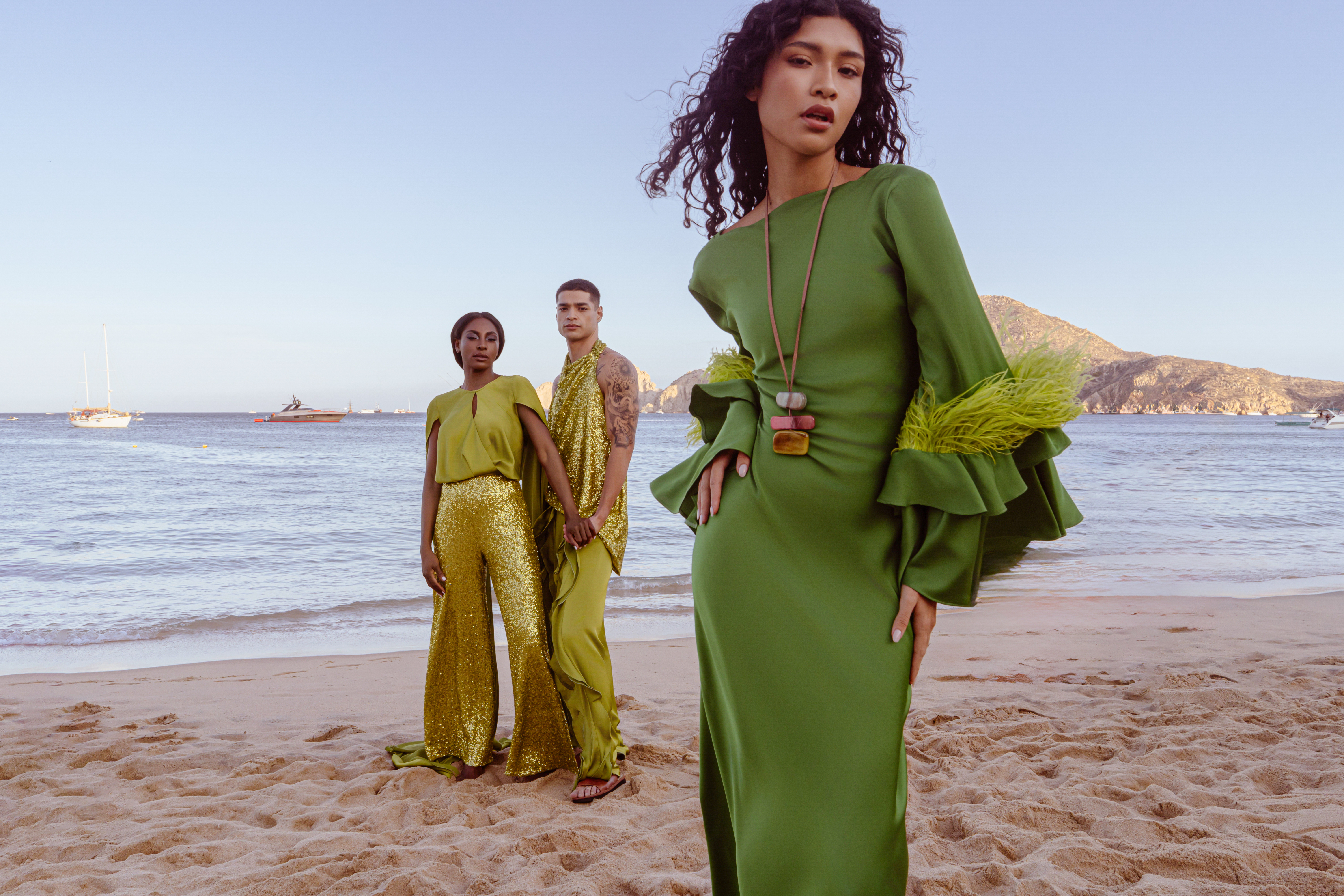 Models in stylish green fashion with sequins, feathers on a sandy beach.