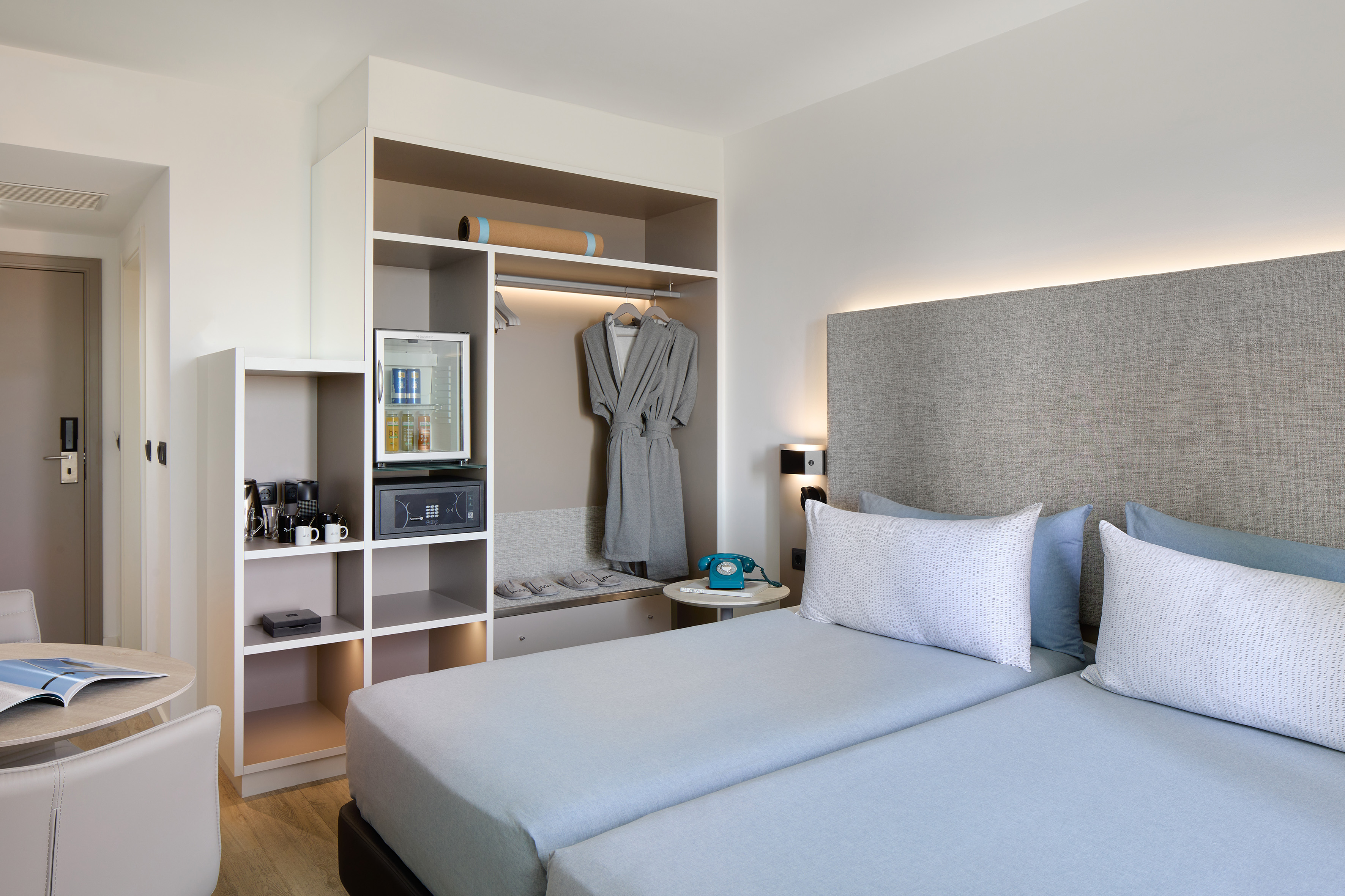 Modern hotel room with twin beds, built-in storage, mini-fridge, and coffee maker.