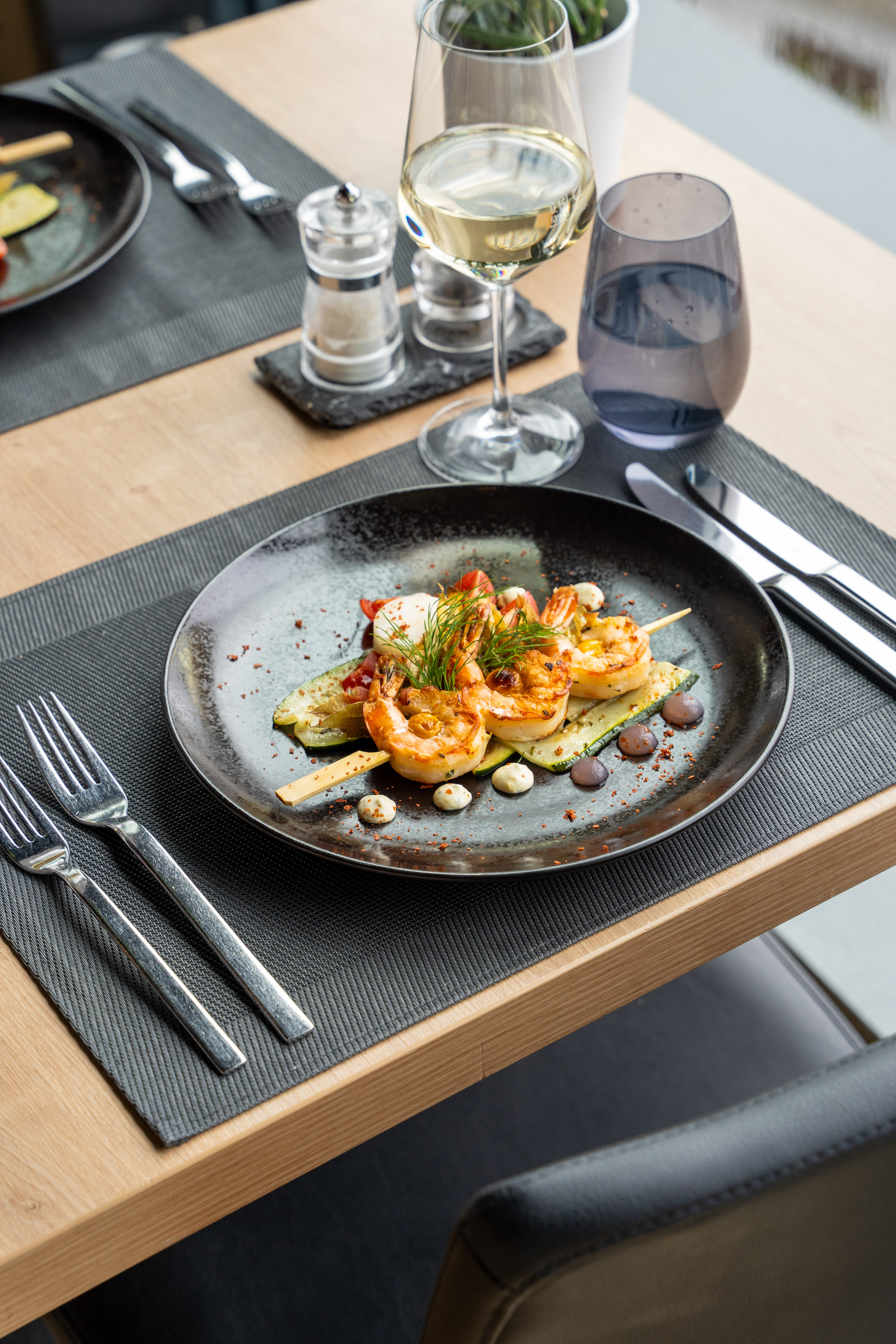 Grilled shrimp skewers with zucchini, sauces, and white wine at a restaurant.