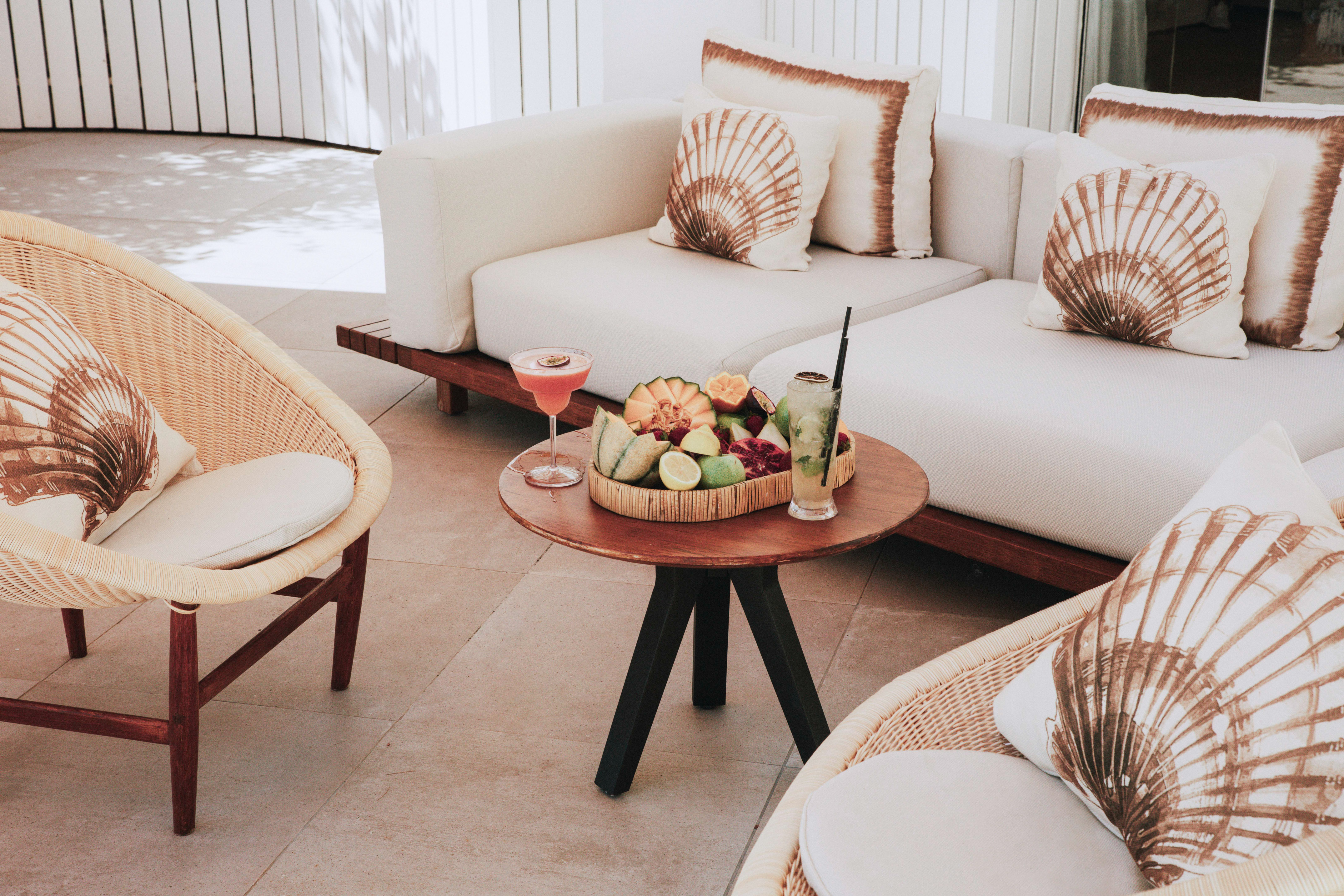 Outdoor lounge with white sofa, wicker chairs, shell pillows, cocktails, and fruit platter.