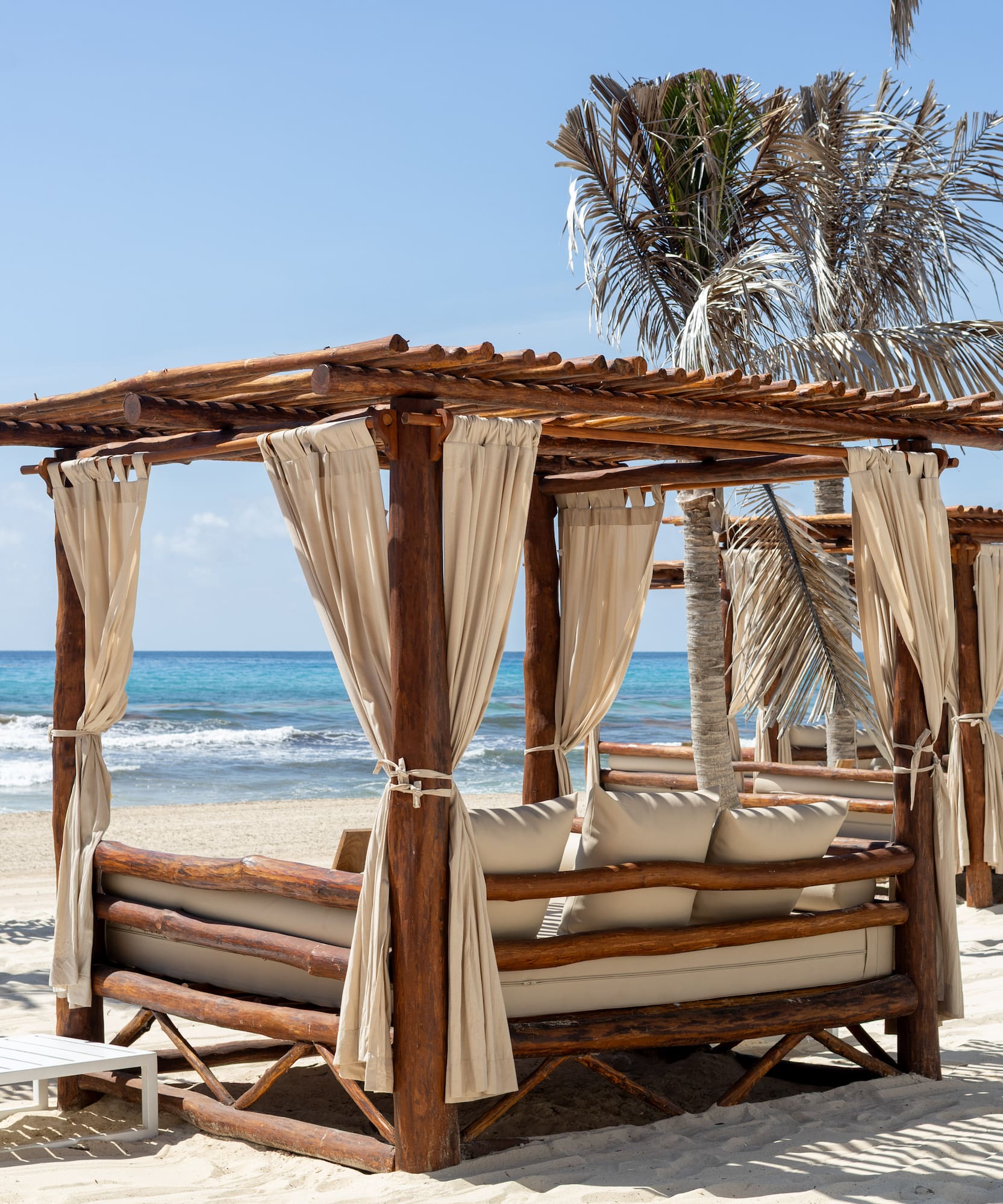 Wooden beach cabanas with cream curtains on white sand, blue ocean.