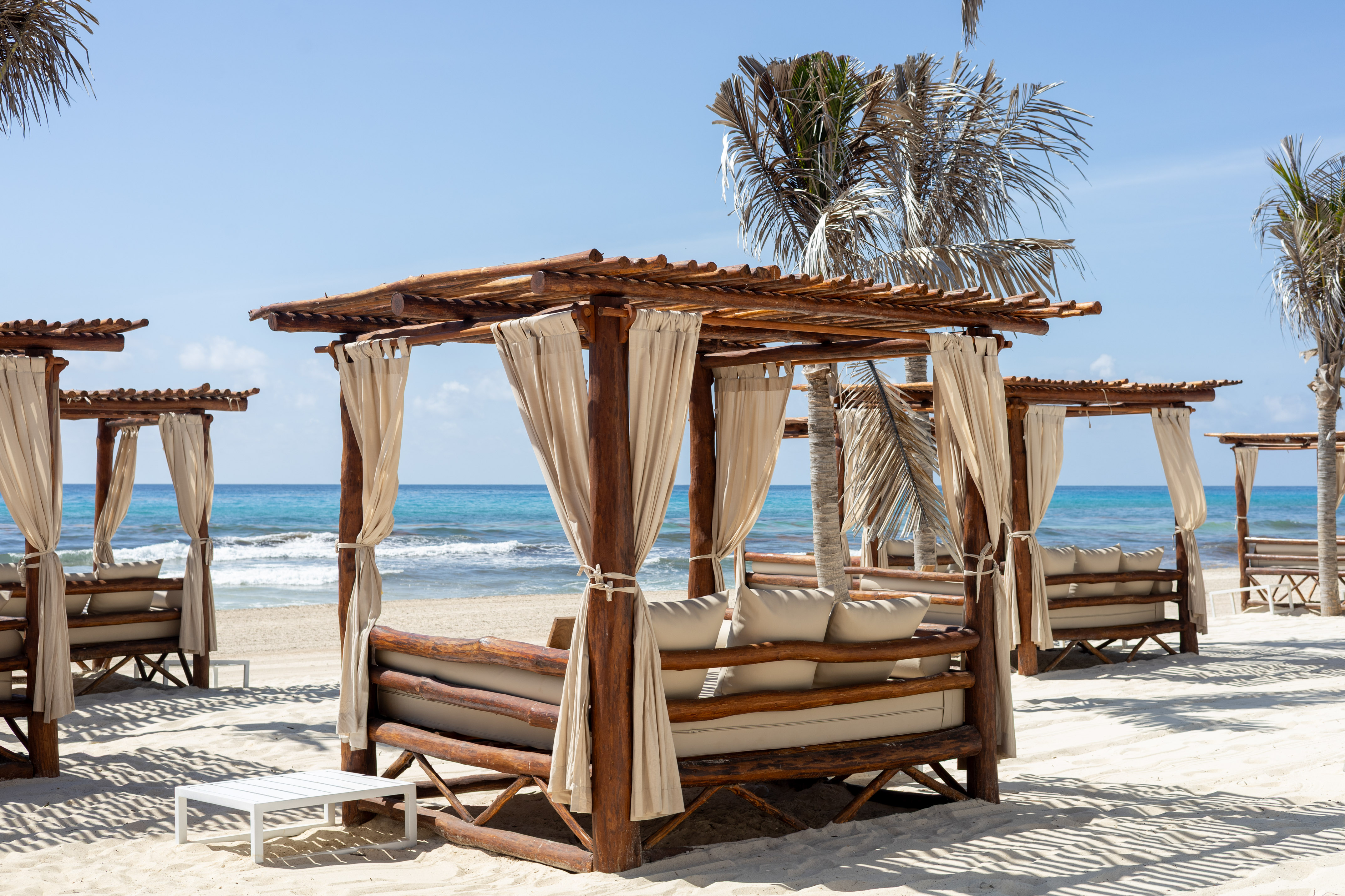 Wooden beach cabanas with cream curtains on white sand, blue ocean.