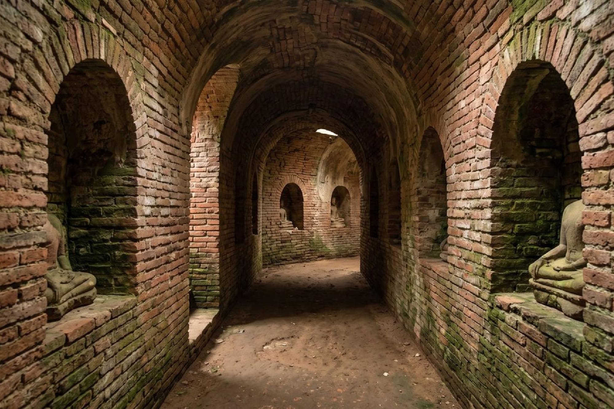 Ancient brick temple corridor with Buddha statues in arched niches.