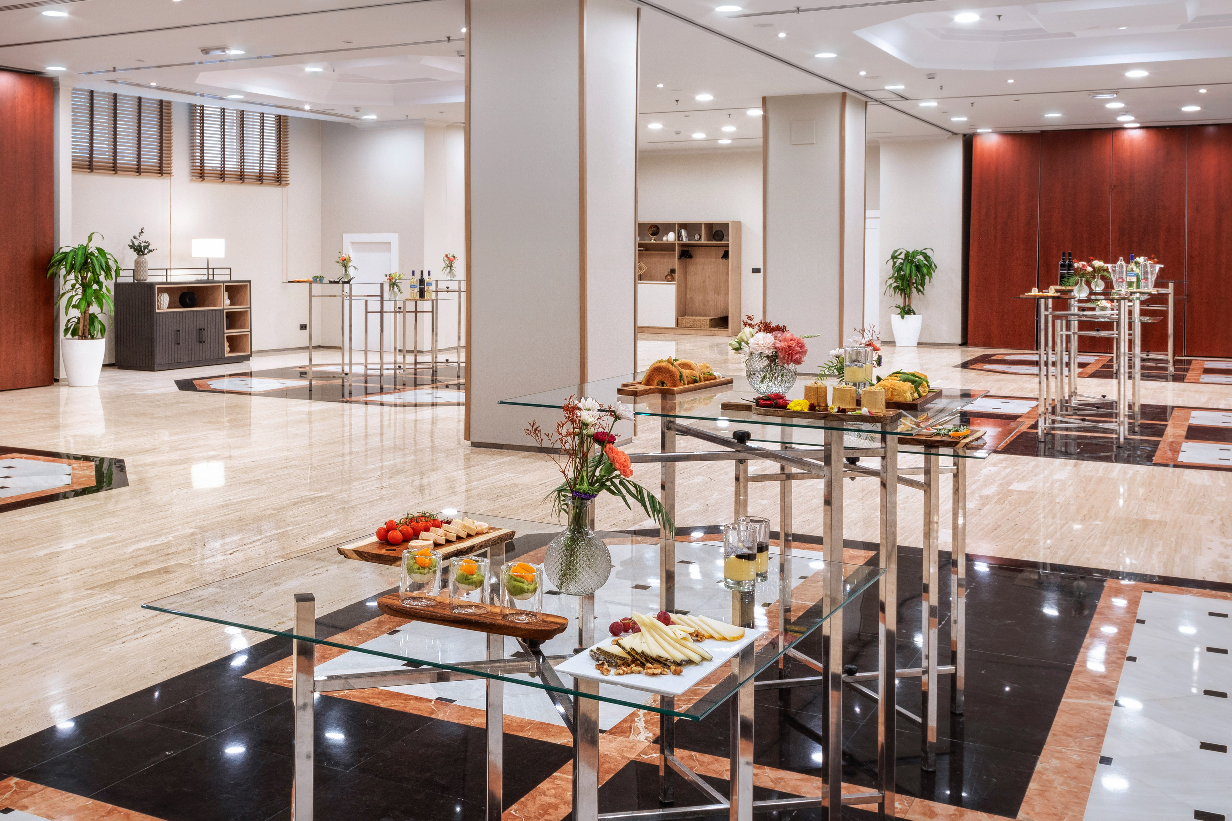 Modern hotel reception with glass tables, buffet snacks, drinks, and flowers.