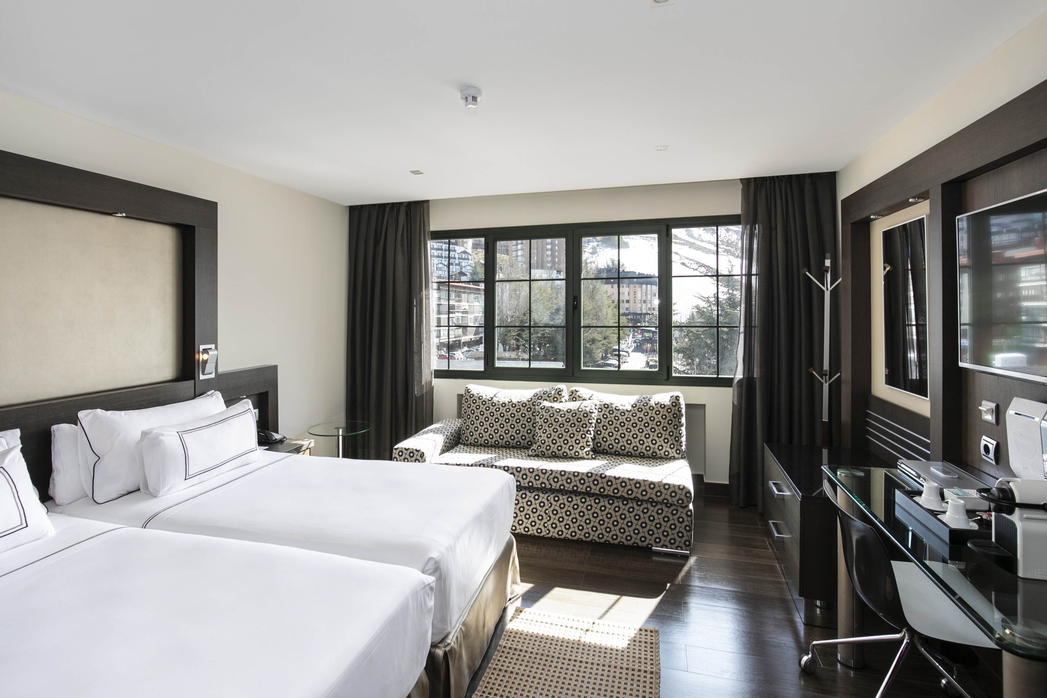 Modern hotel room with twin beds, sofa, work desk, and scenic mountain view.