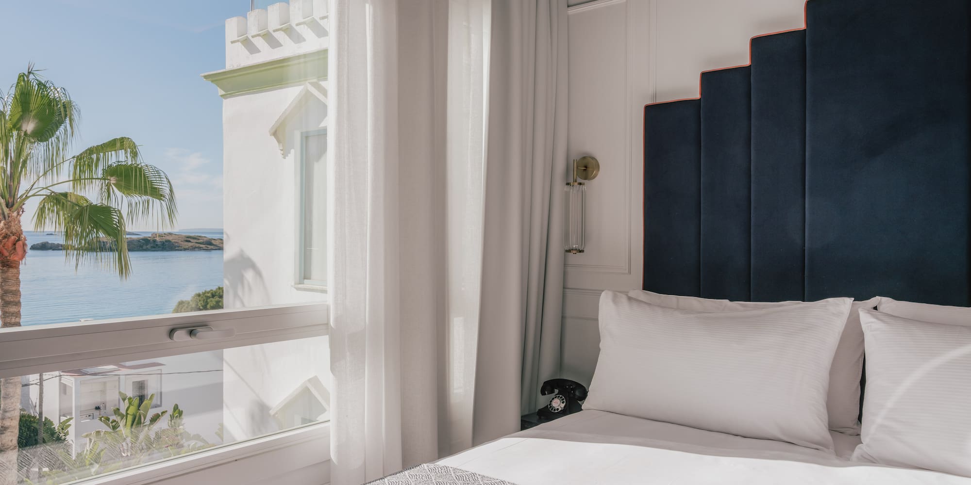 Coastal hotel bedroom with sea view, palm tree, and elegant stepped headboard.