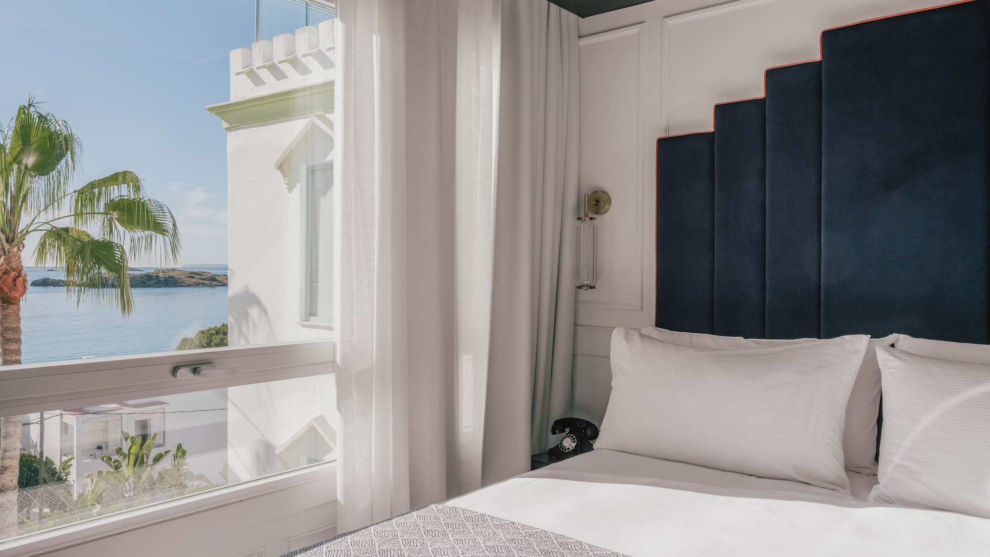 Coastal hotel bedroom with sea view, palm tree, and elegant stepped headboard.
