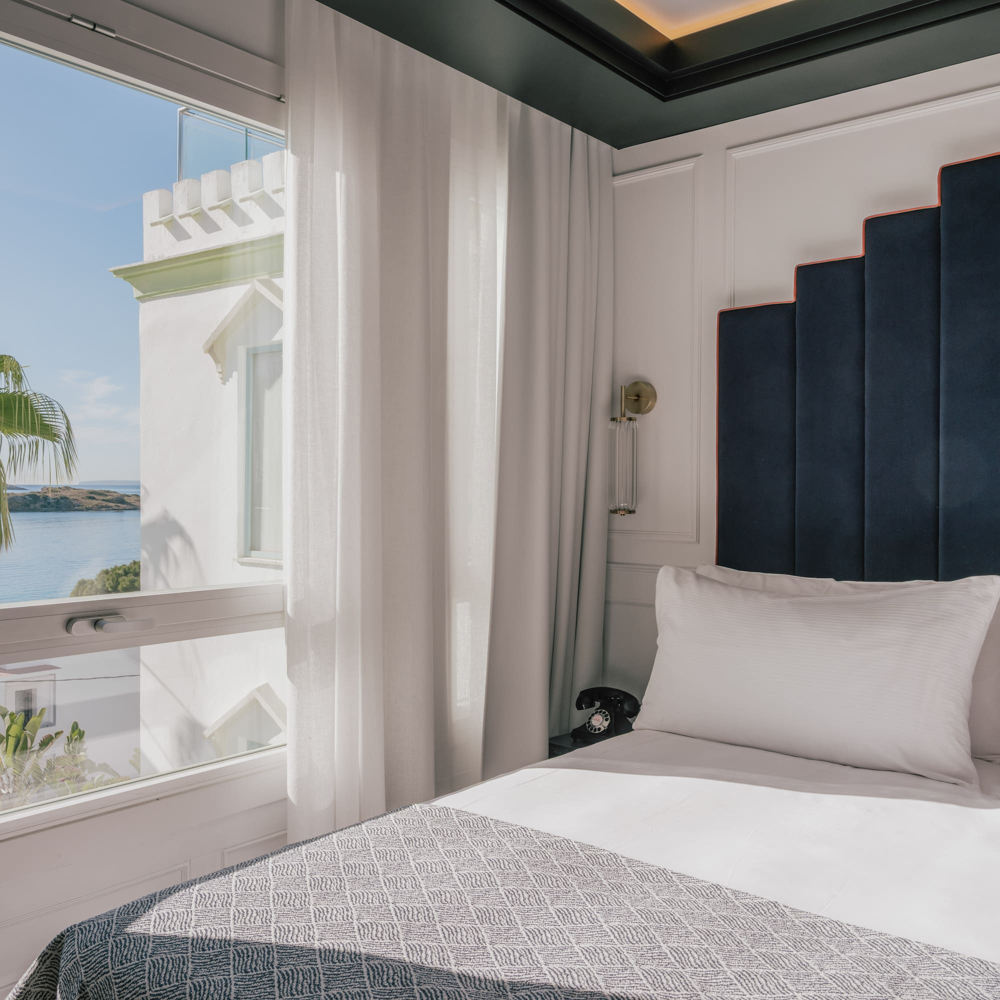 Coastal hotel bedroom with sea view, palm tree, and elegant stepped headboard.