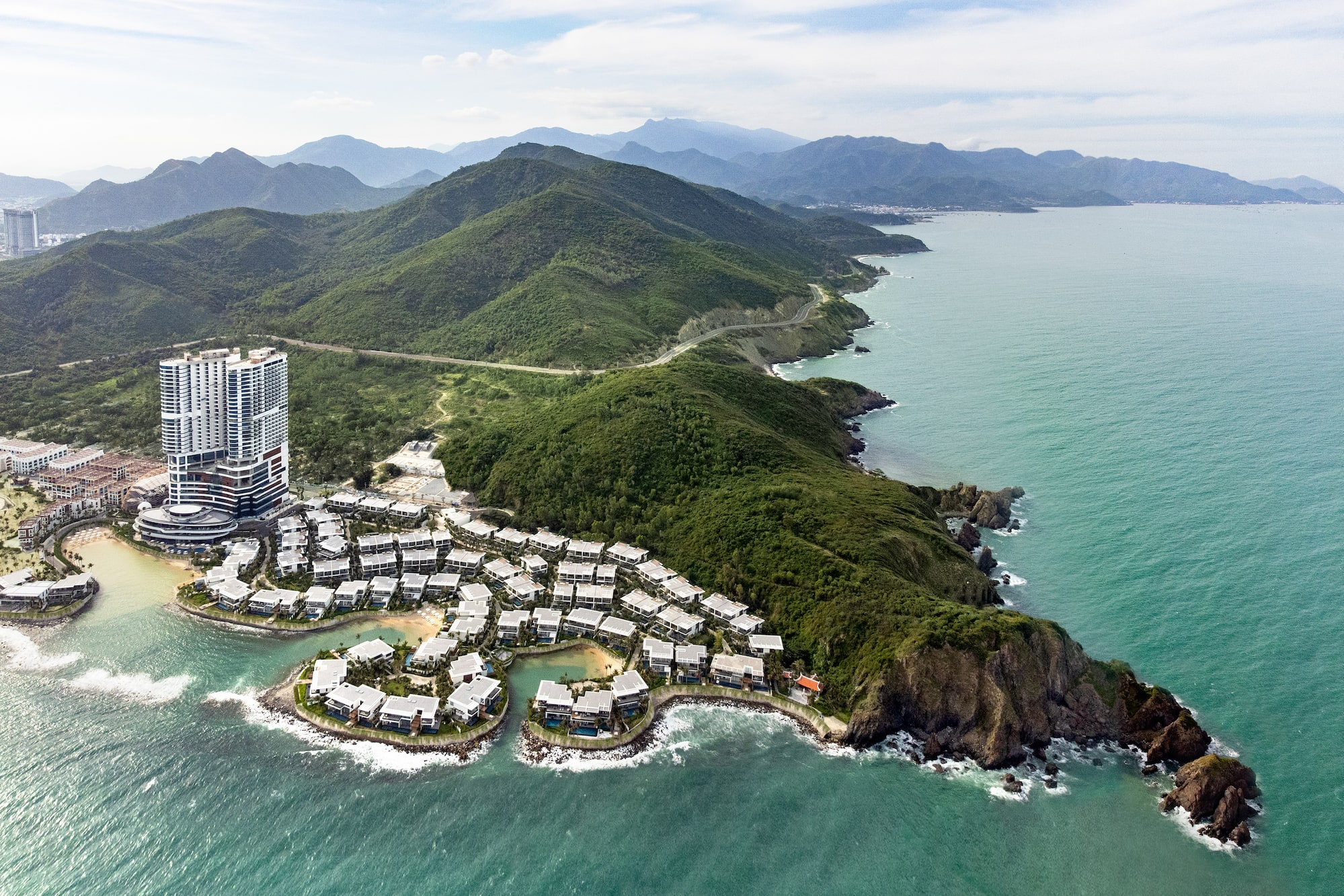 Aerial view: Oceanfront resort with high-rise, villas, green mountains, turquoise sea.