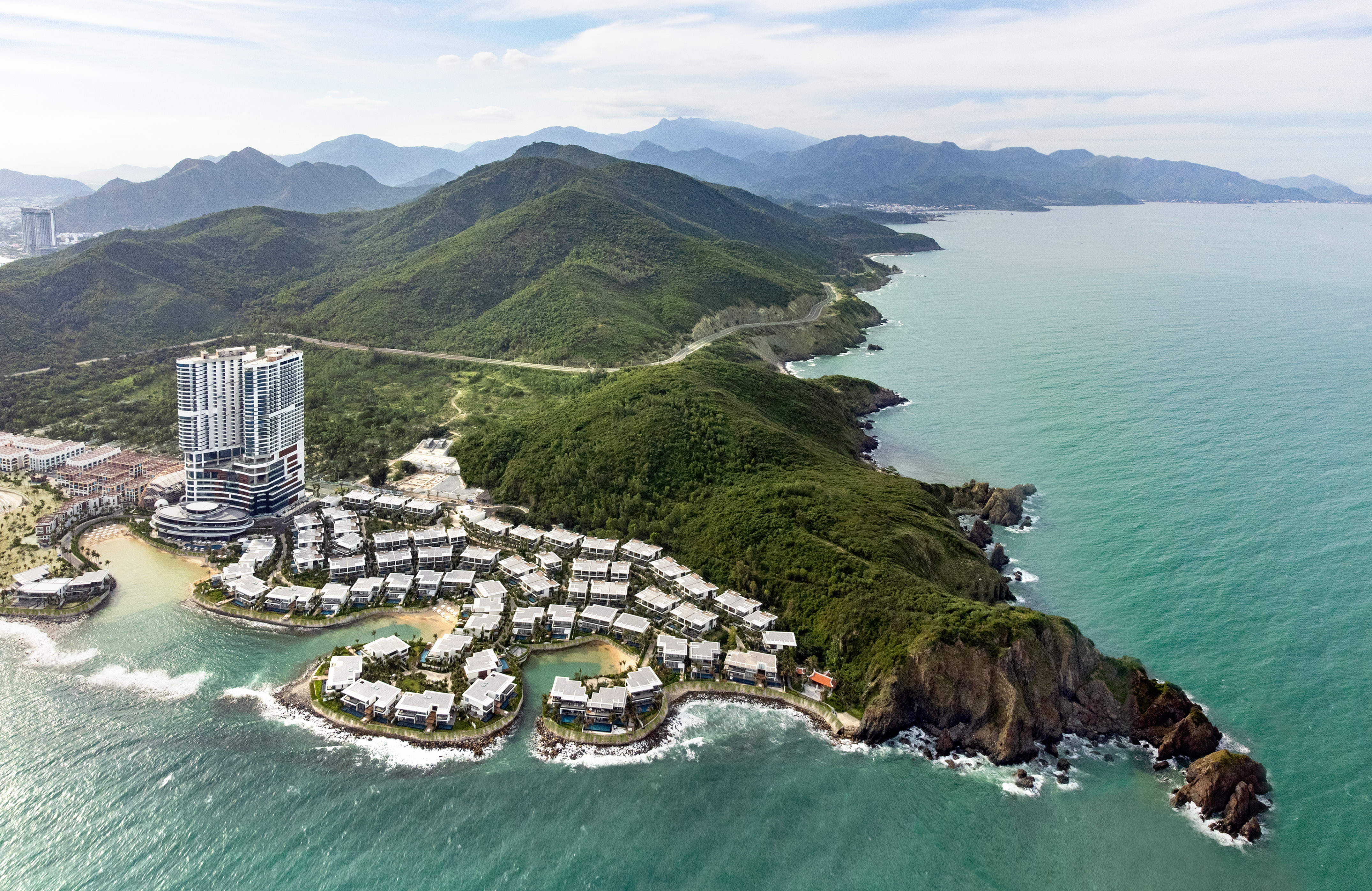 Aerial view: Oceanfront resort with high-rise, villas, green mountains, turquoise sea.