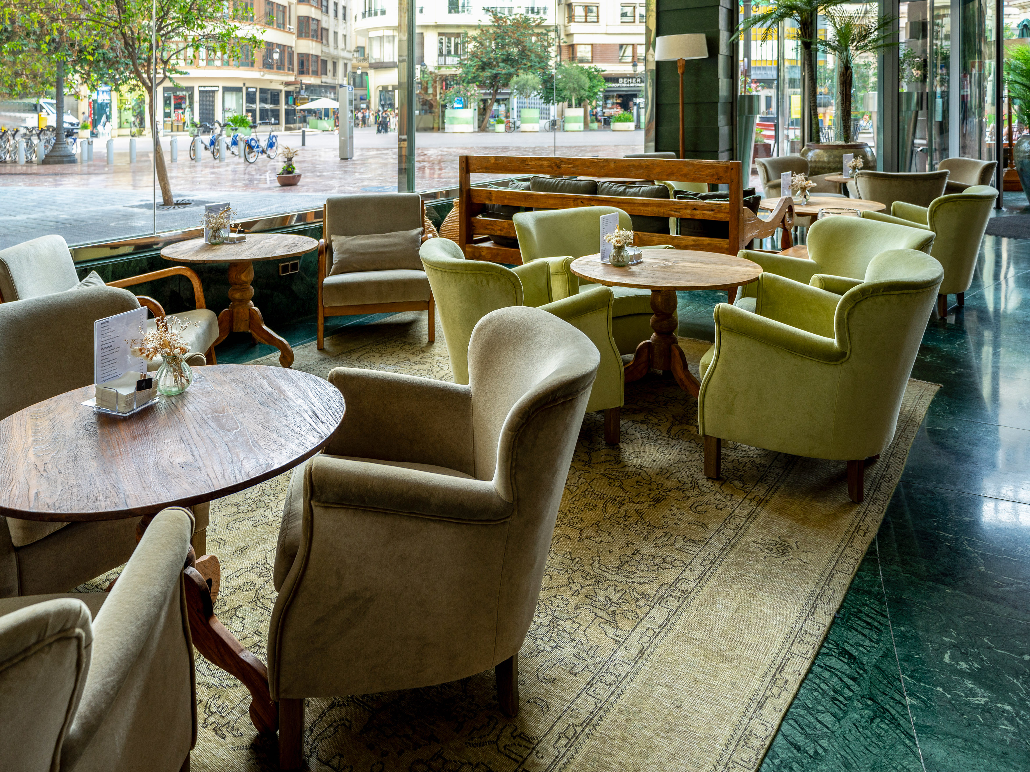 Cafe lounge with green and beige armchairs, wooden tables, and city street view.