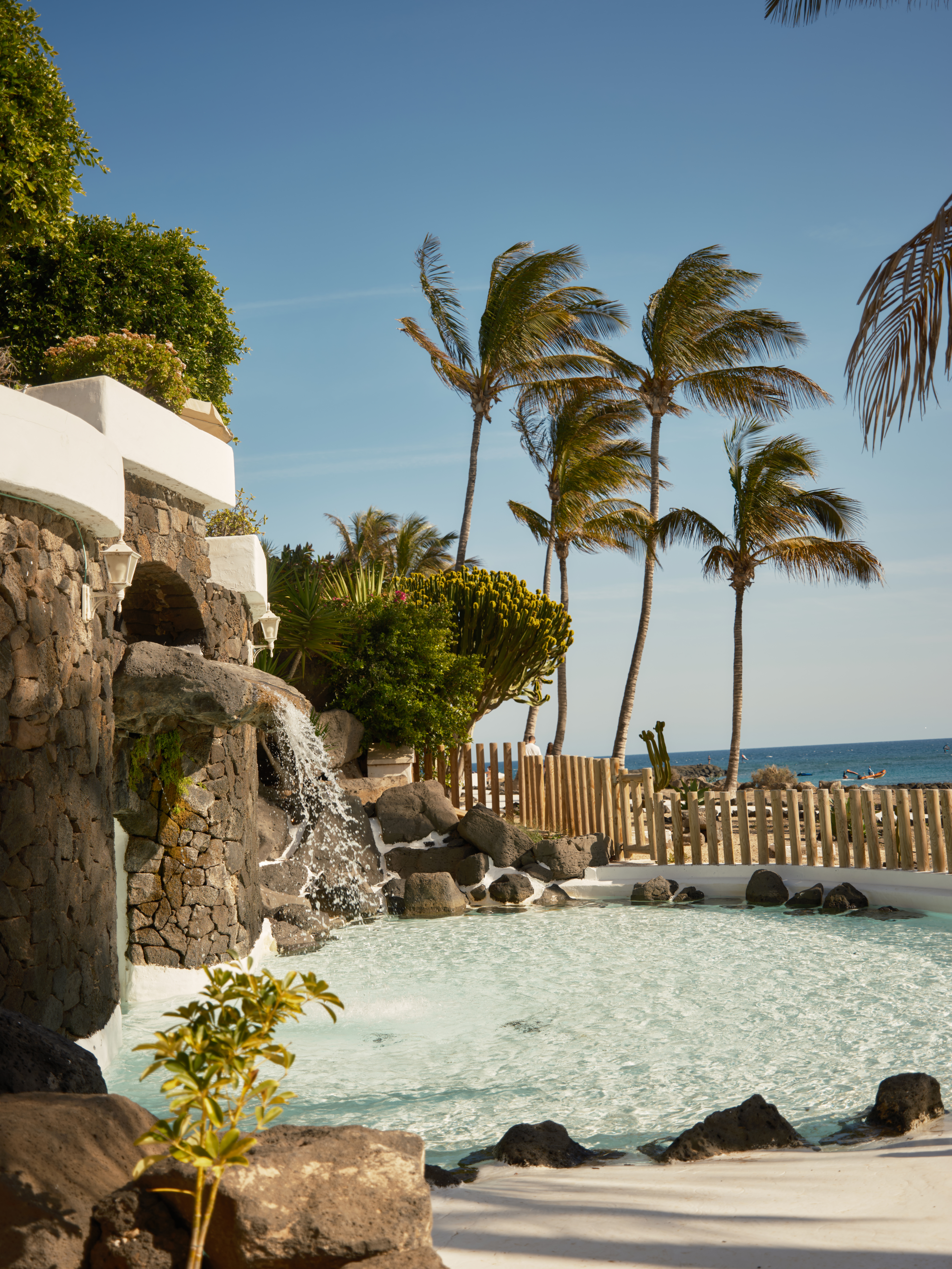 Piscina resort con cascada volcánica, palmeras y mar. Destino tropical.