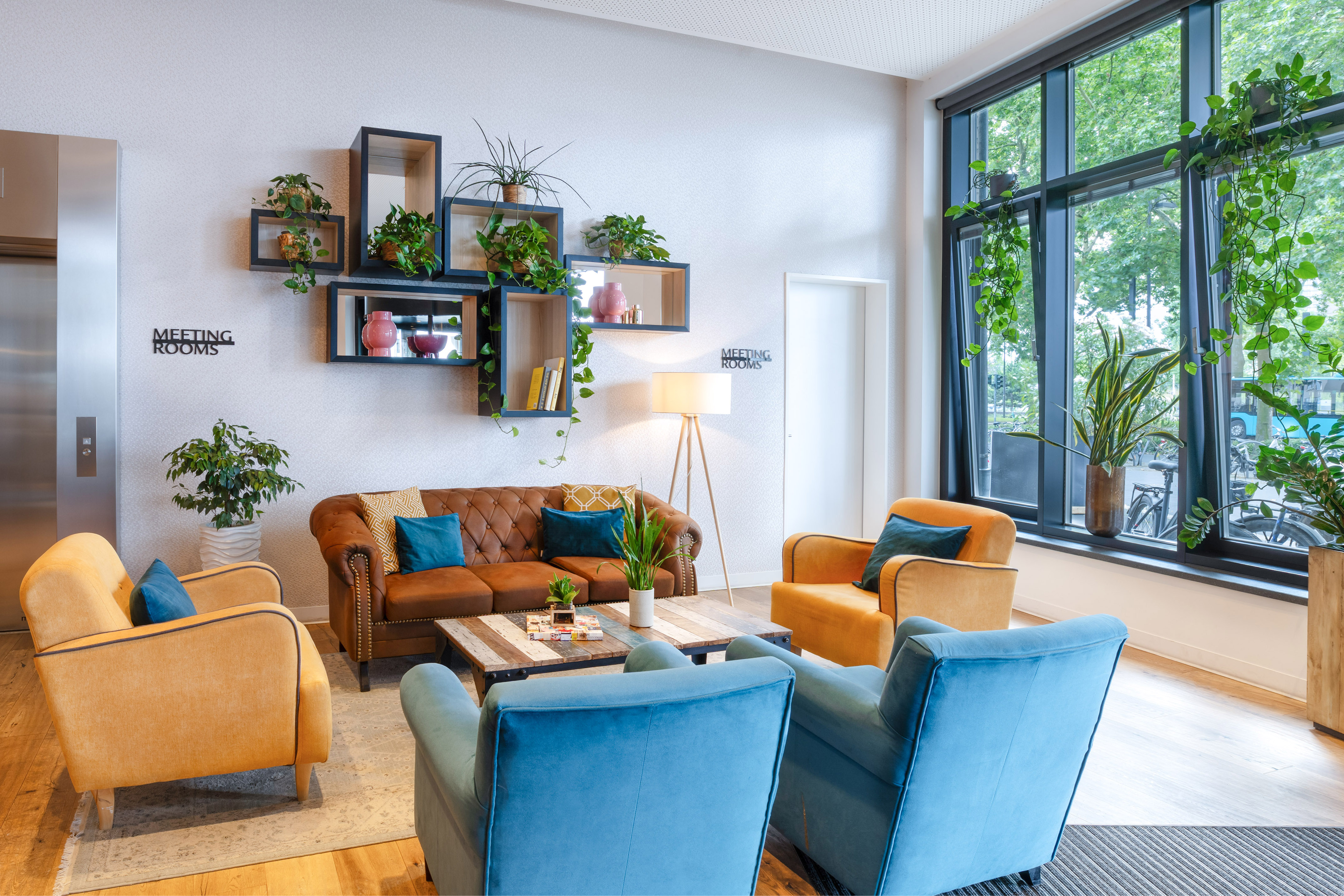 Cozy hotel lobby lounge with colorful seating, abundant plants, and meeting rooms sign.