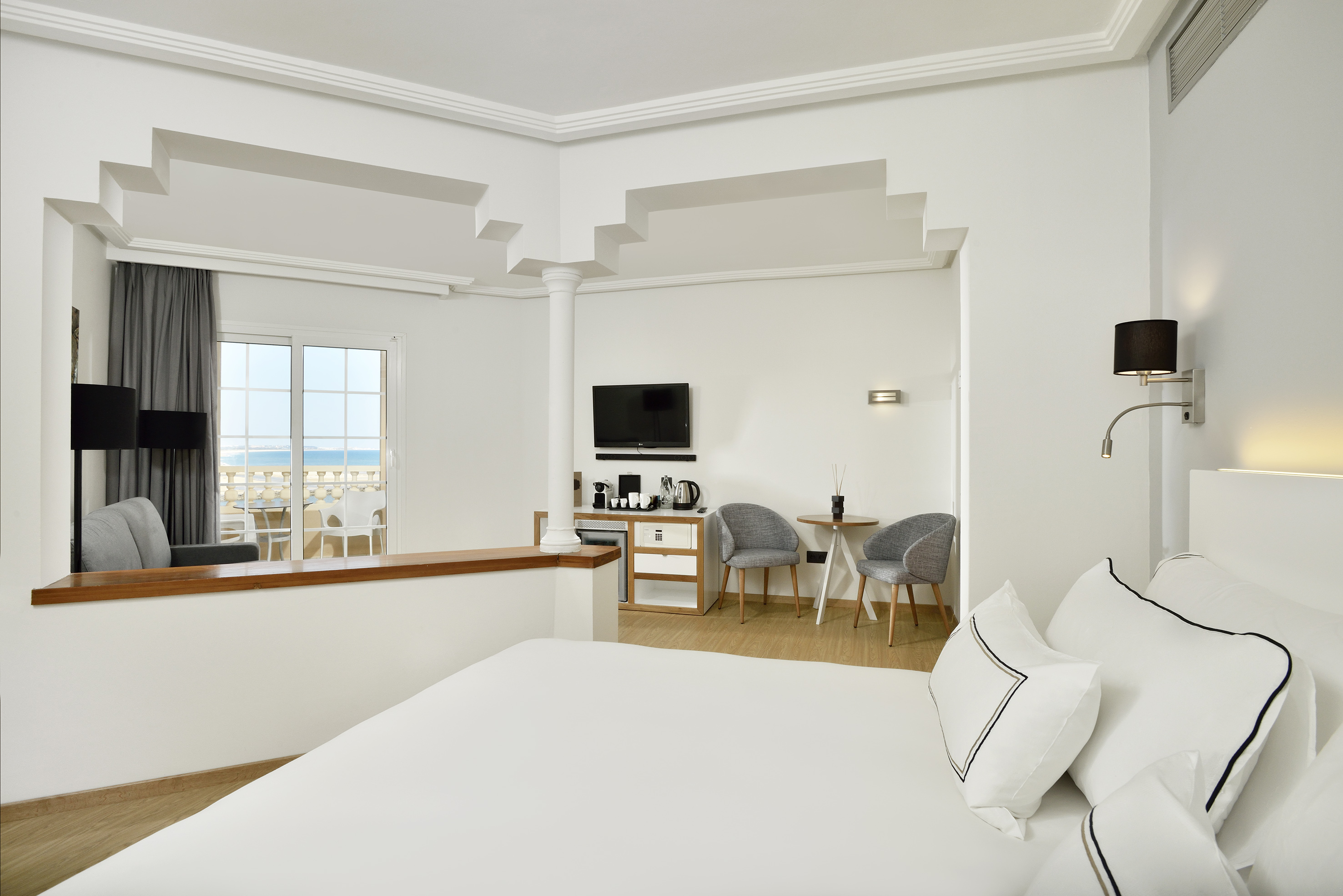 Modern hotel suite with sea view balcony, white bedroom and sitting area.