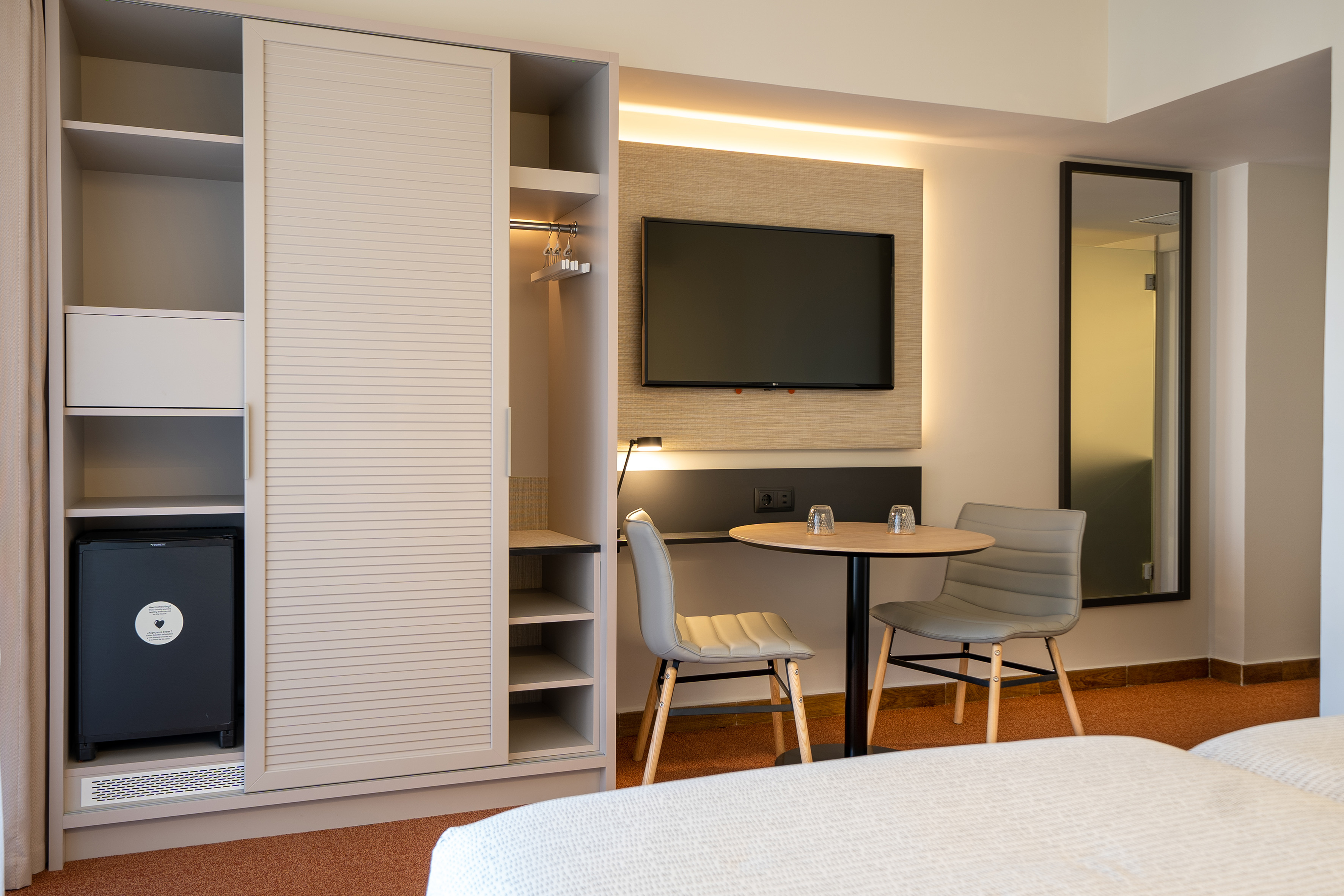 Modern hotel room with wardrobe, minibar, wall-mounted TV, and table with two chairs.