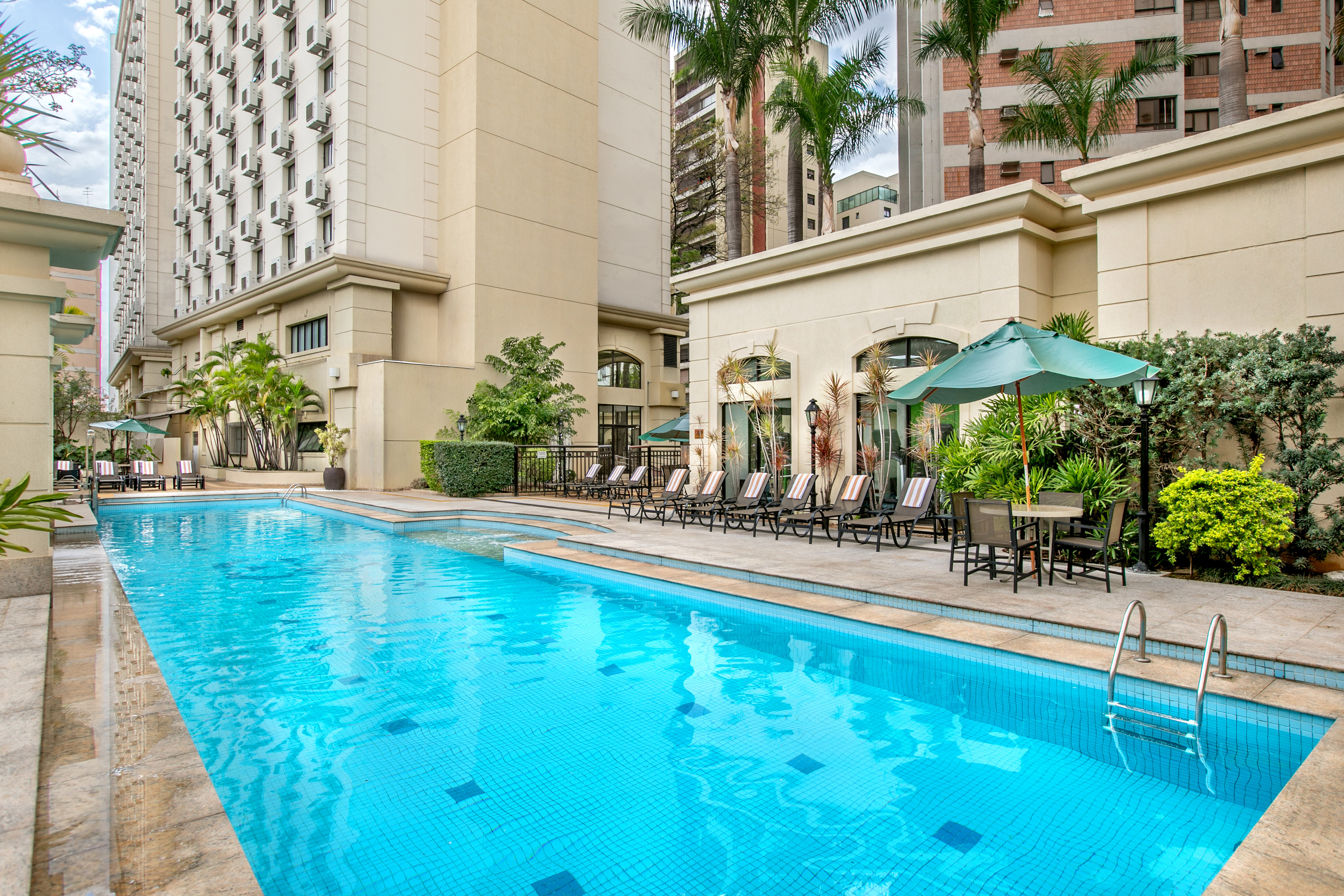 Bright blue hotel pool with lounge chairs, umbrella, and tropical palm trees.