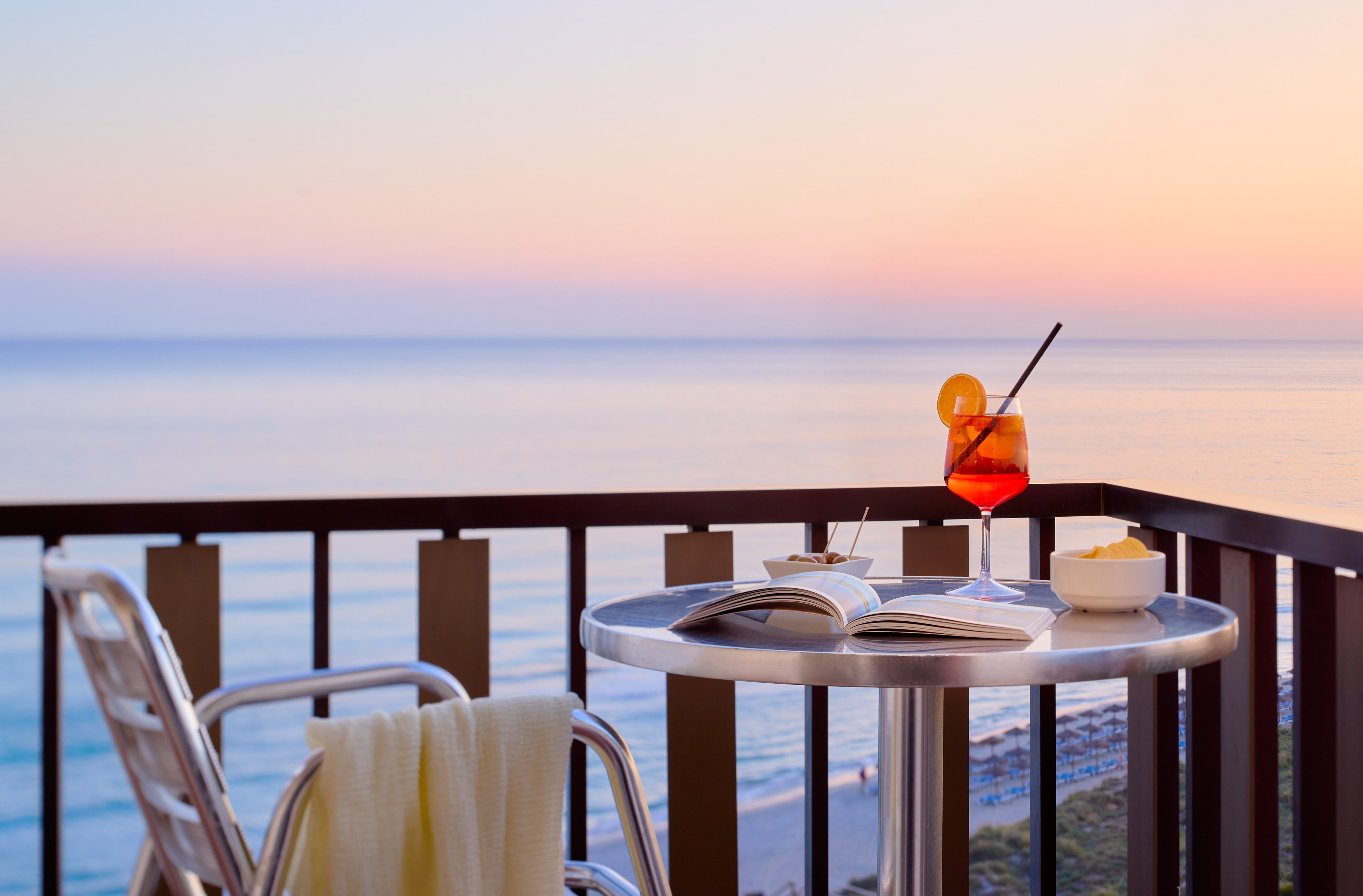 Balcony with cocktail, open book, snacks, and sunset ocean view.