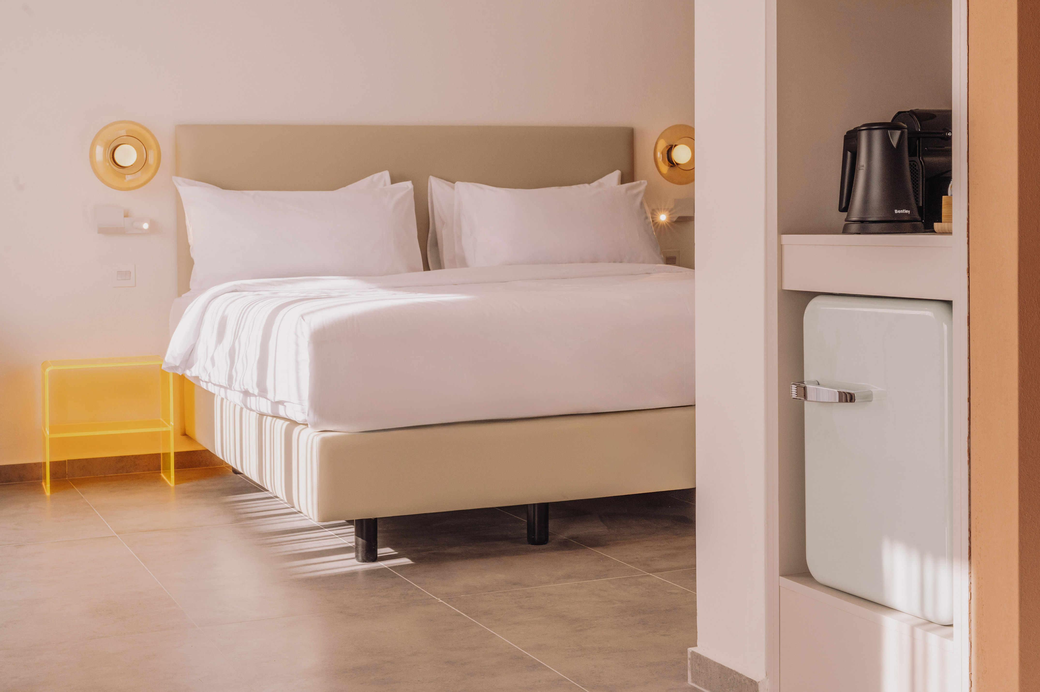 Minimalist hotel bedroom featuring white bed, beige headboard, and mini-bar.