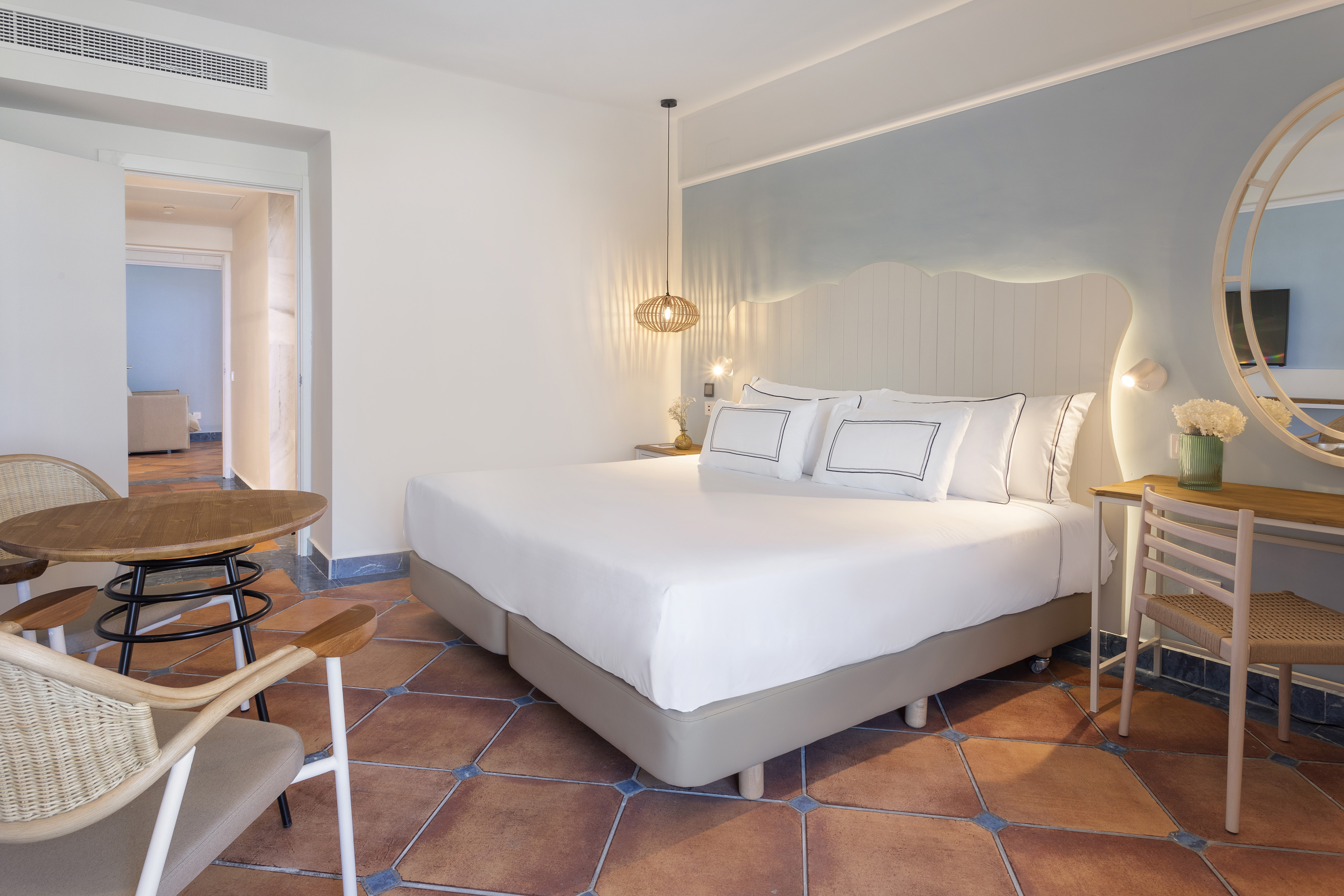 Stylish hotel room with white bed, terracotta floor, and rattan furniture.