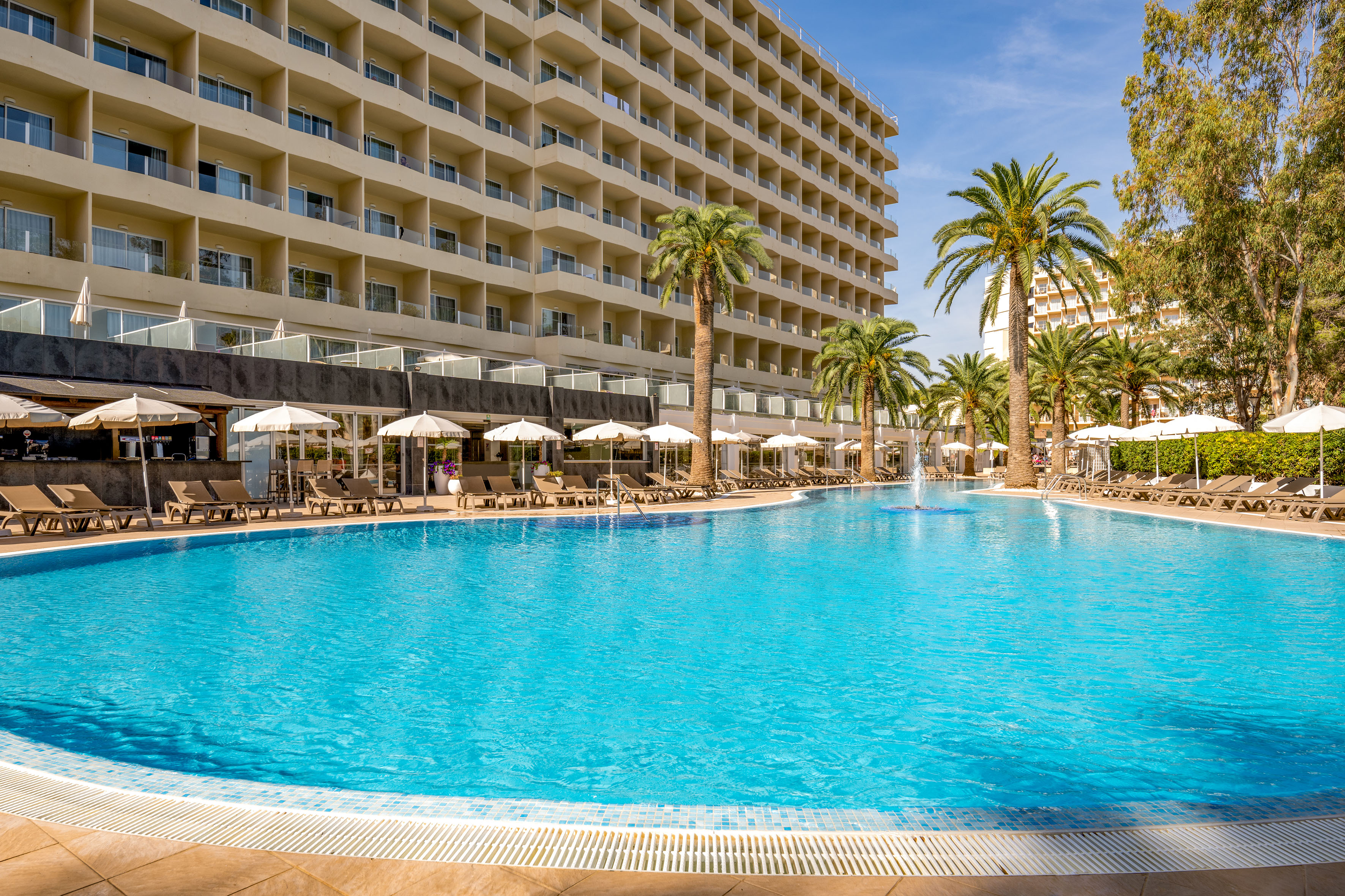 Hotel resort with large swimming pool, palm trees, sun loungers, and poolside bar.