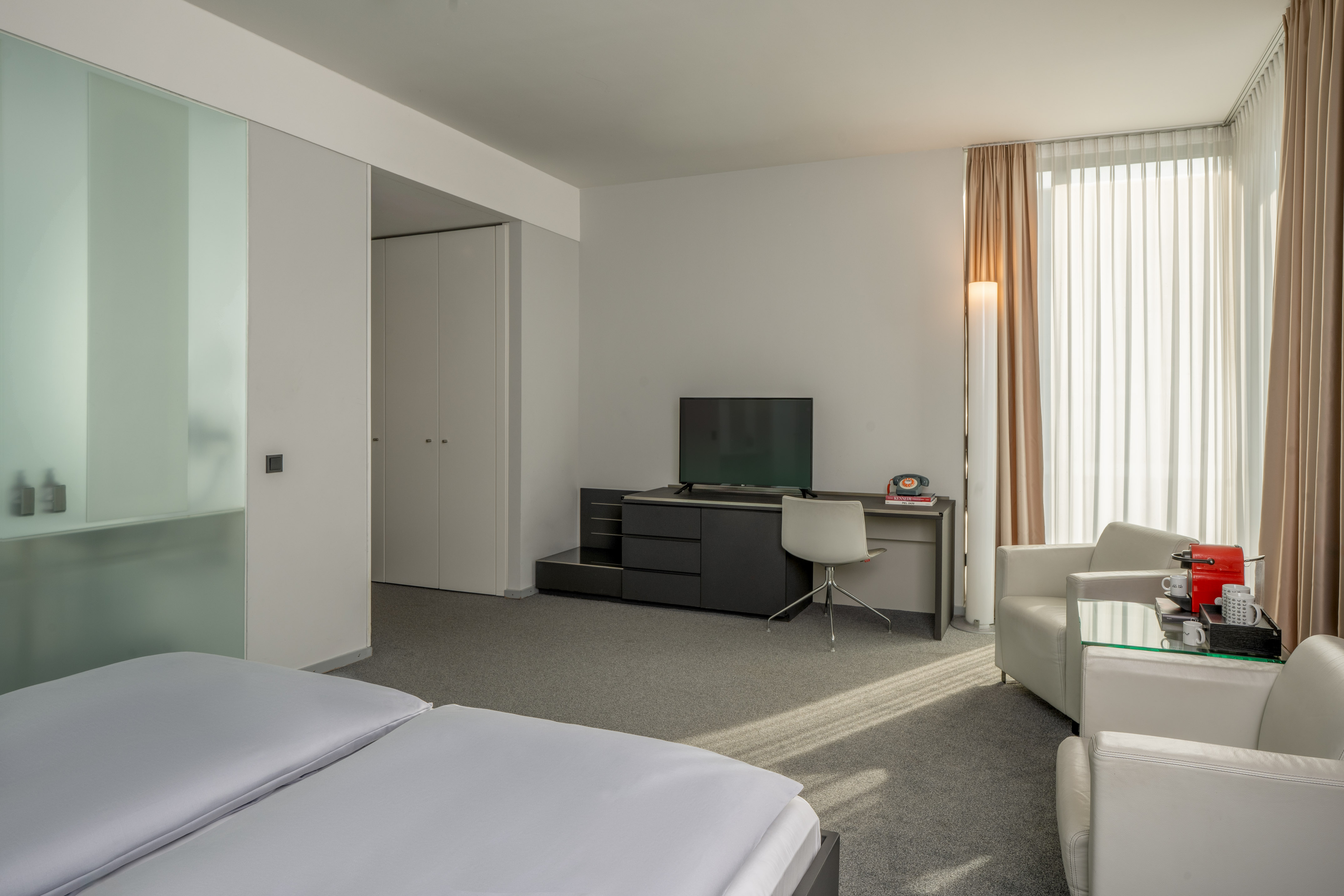 Modern hotel room with twin beds, desk, TV, armchairs, and glass partition.