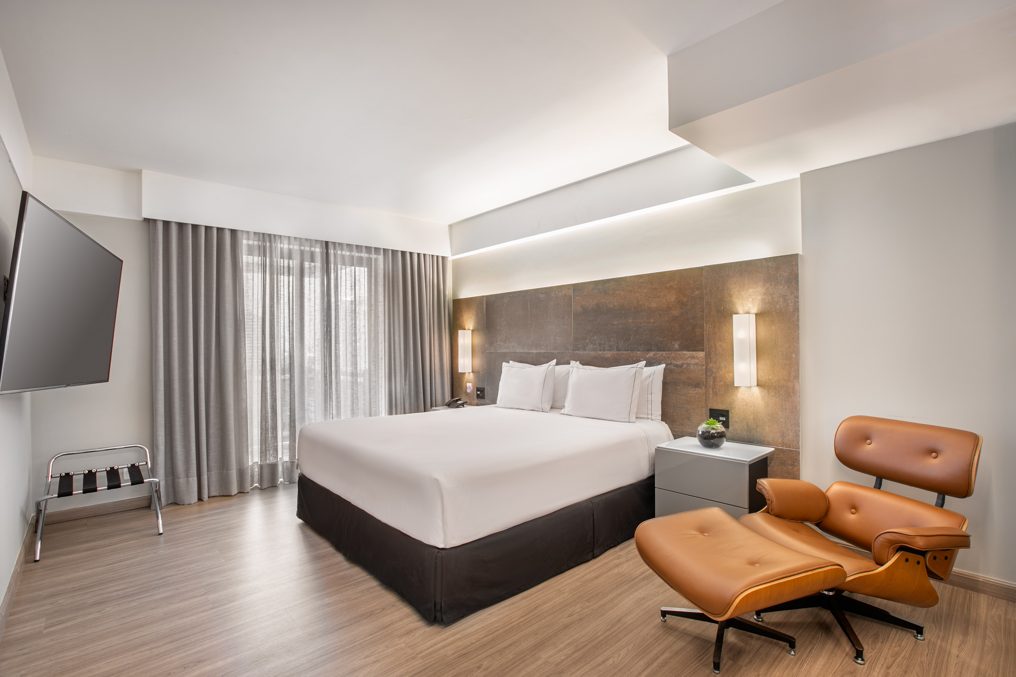 Modern hotel bedroom with king bed, brown leather lounge chair, and TV.
