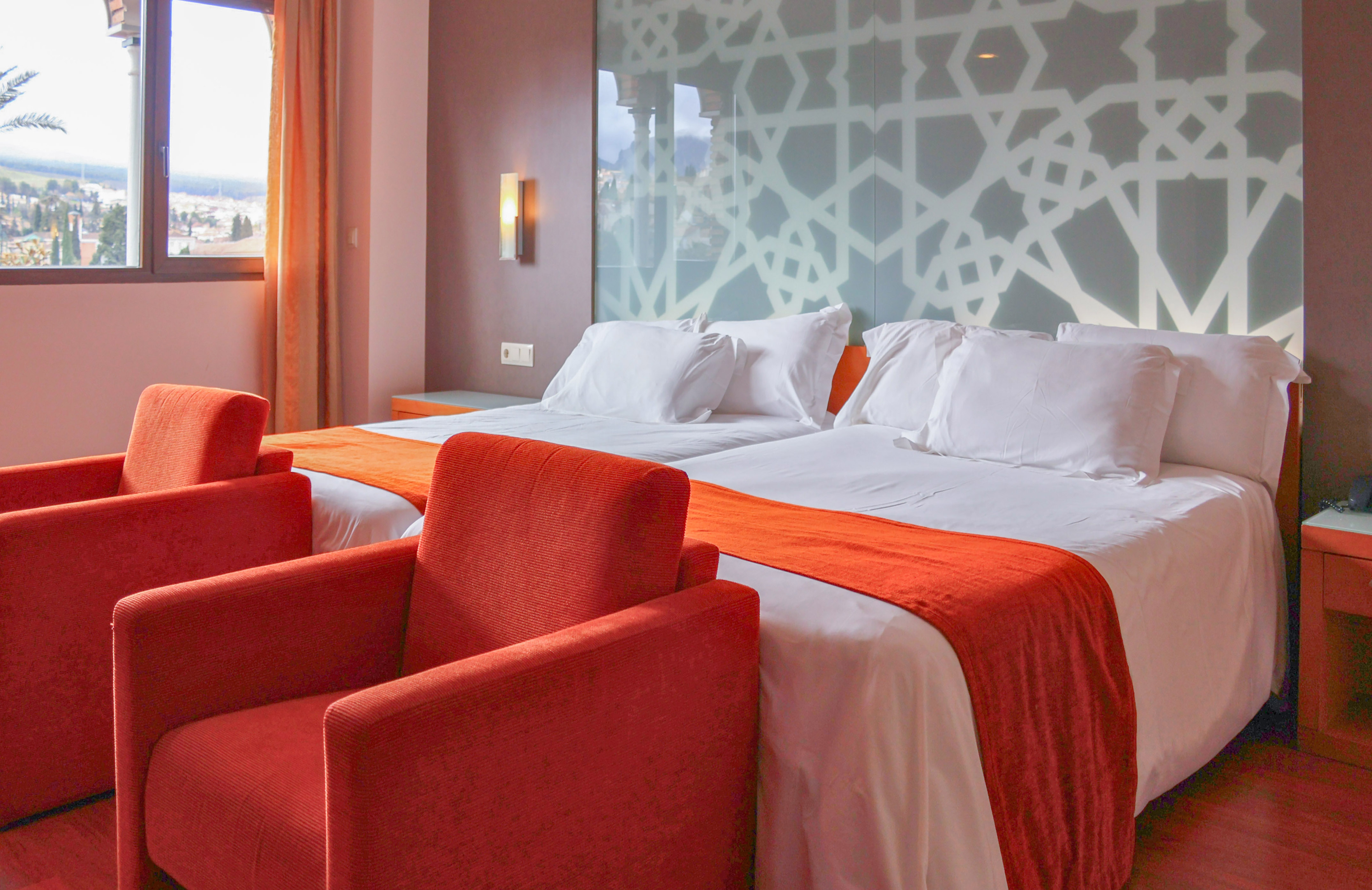 Hotel room with twin beds, red armchairs, geometric headboard, city view.