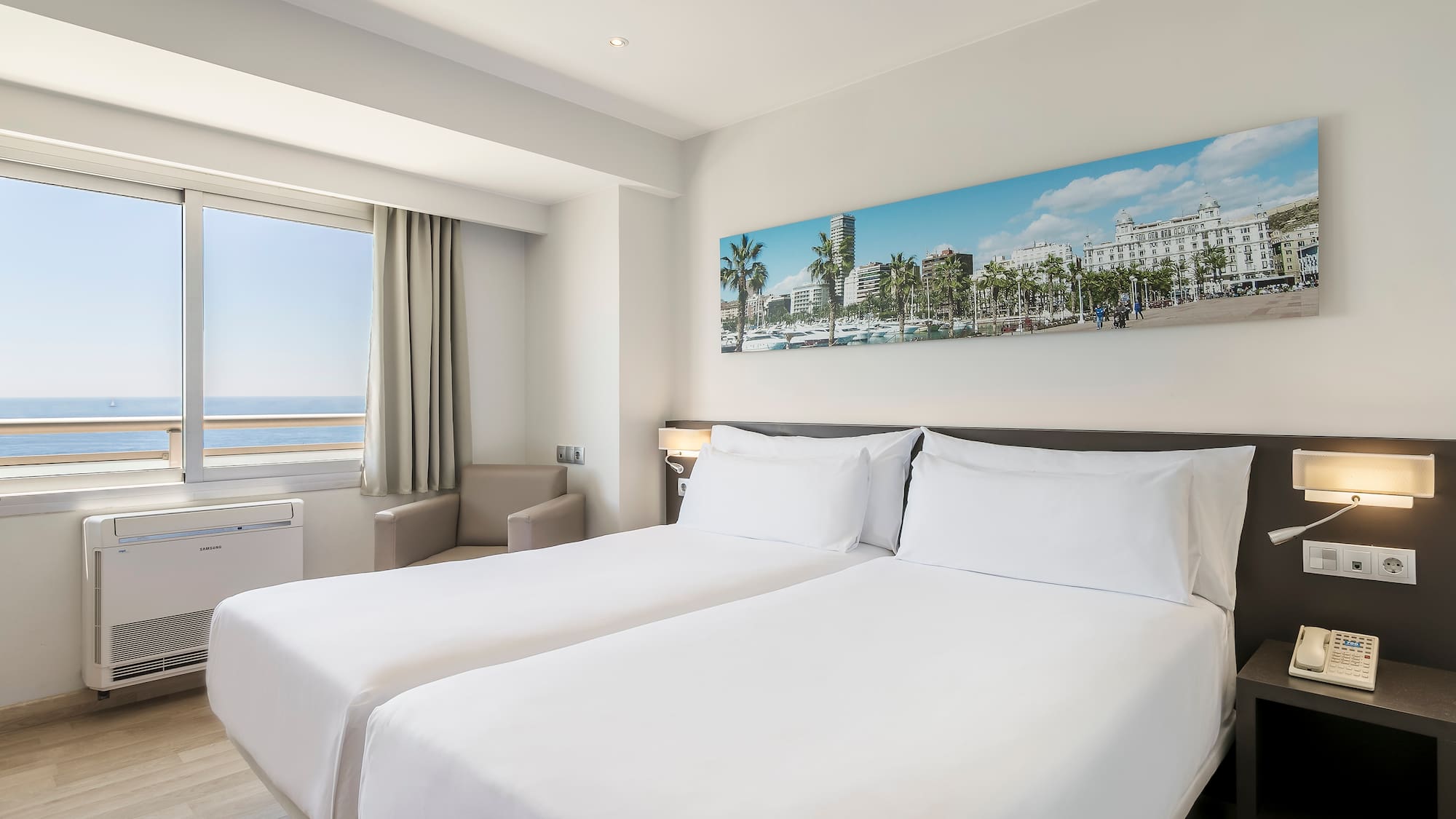 Modern hotel room with ocean view, twin beds, and cityscape artwork.
