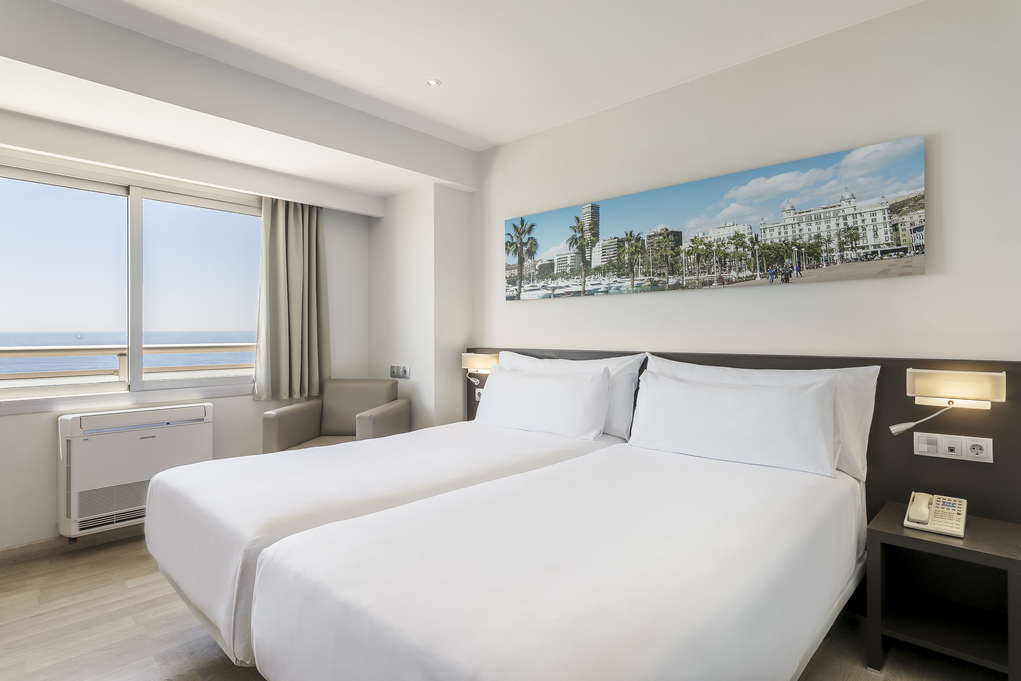 Modern hotel room with ocean view, twin beds, and cityscape artwork.
