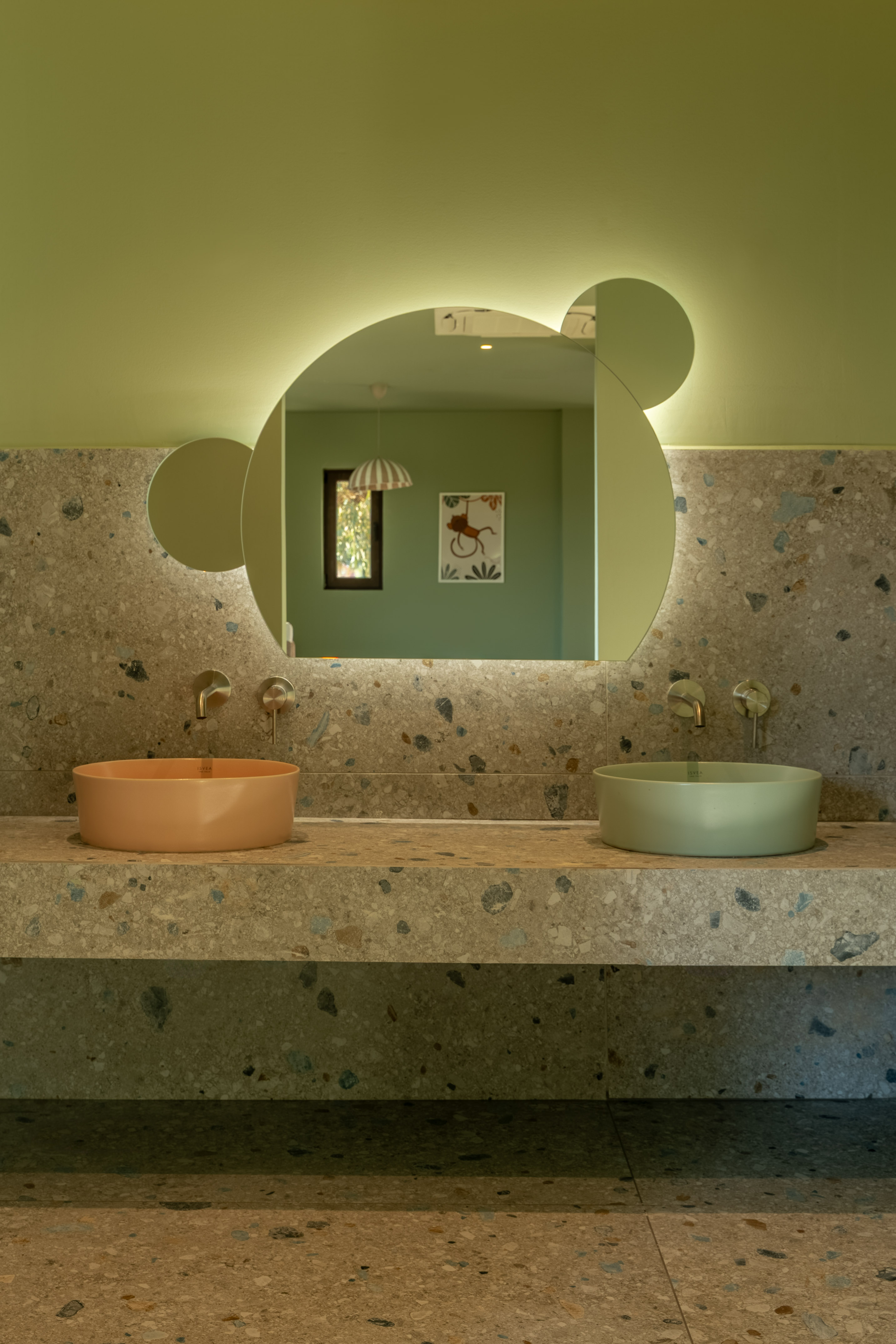 Terrazzo kids bathroom vanity with peach and green sinks, backlit mirror.