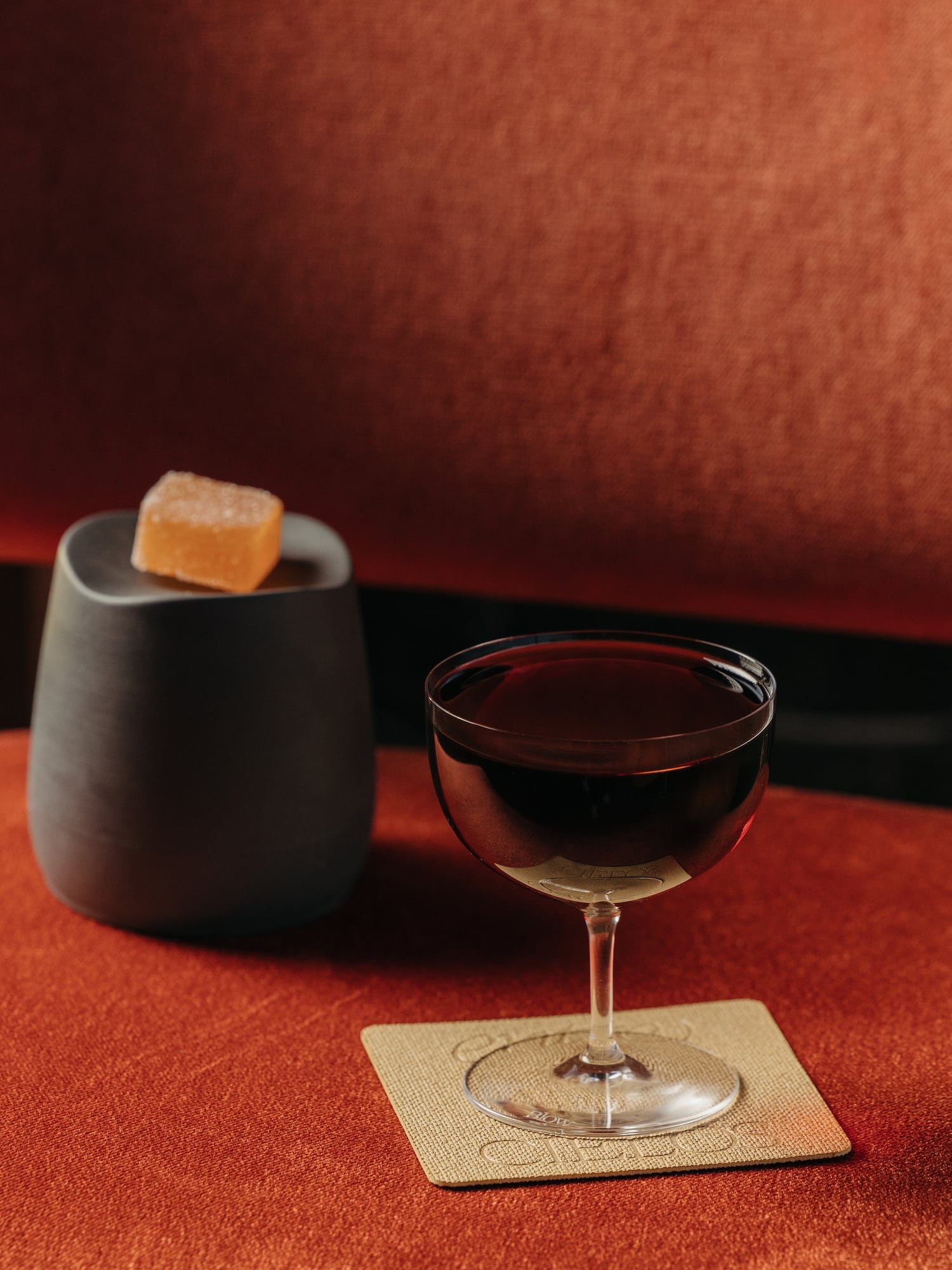 Cóctel oscuro en copa, gominola de naranja, recipiente gris sobre textura roja.