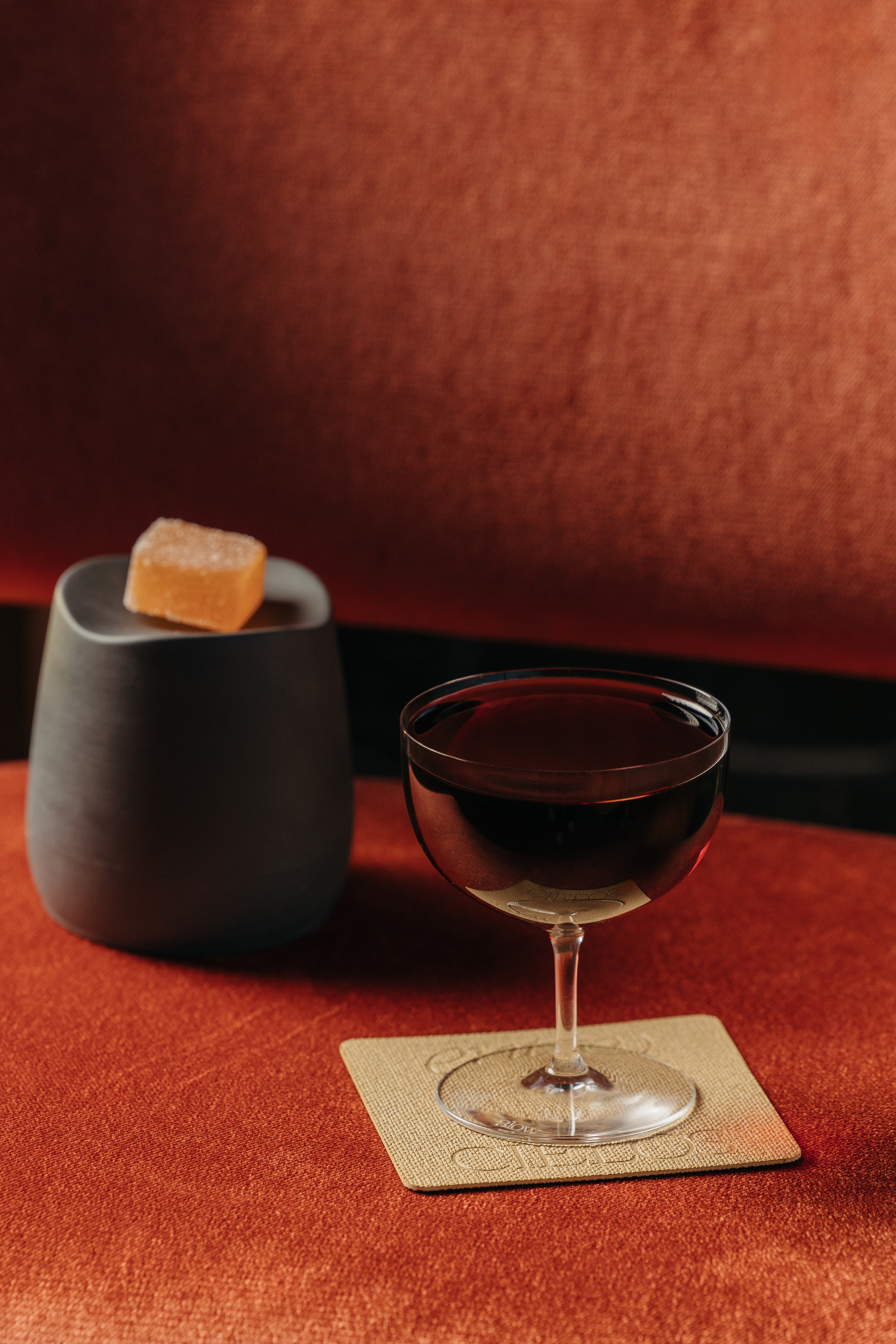 Cóctel oscuro en copa, gominola de naranja, recipiente gris sobre textura roja.