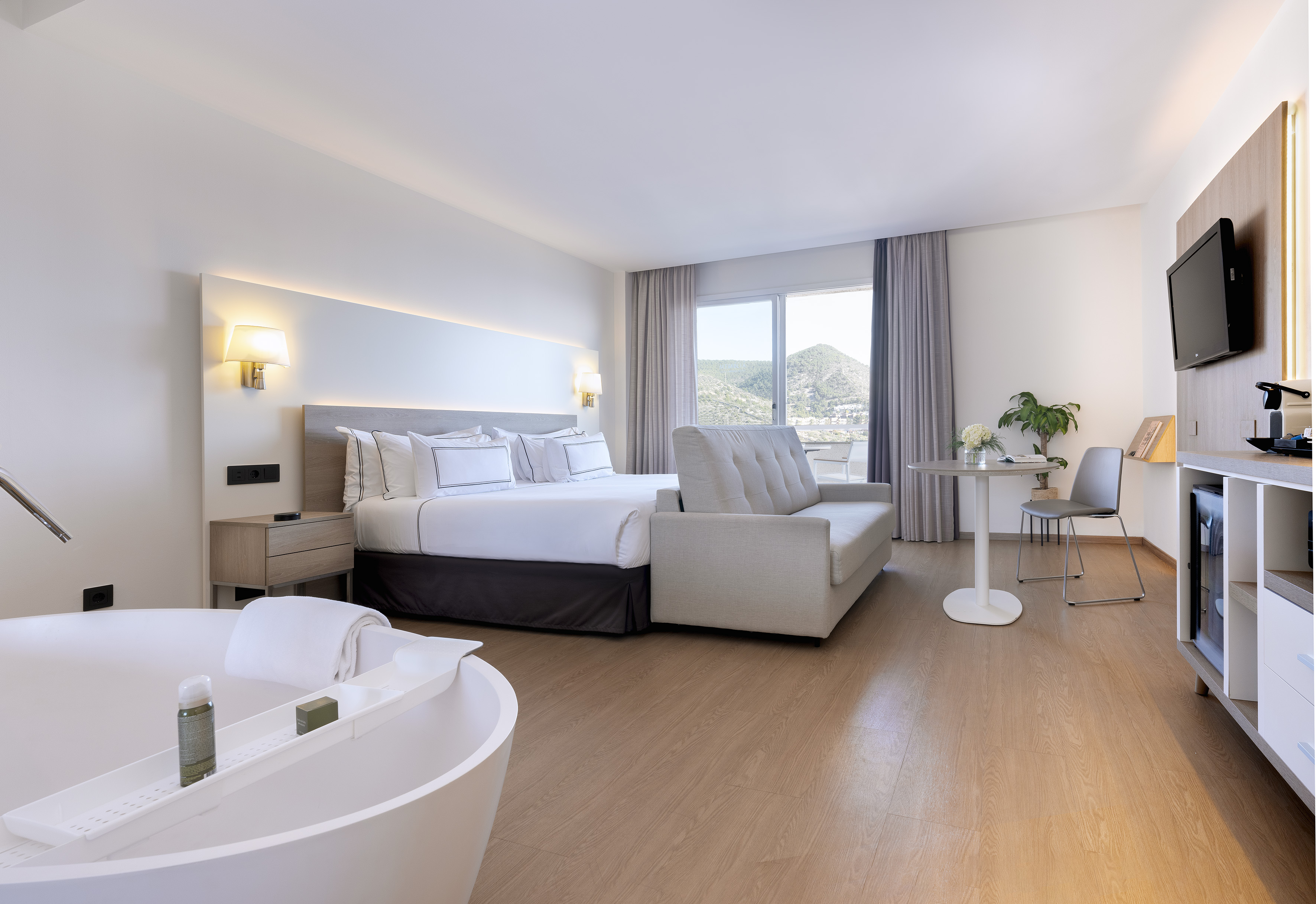 Modern hotel room with king bed, freestanding tub, sofa bed, and mountain view.