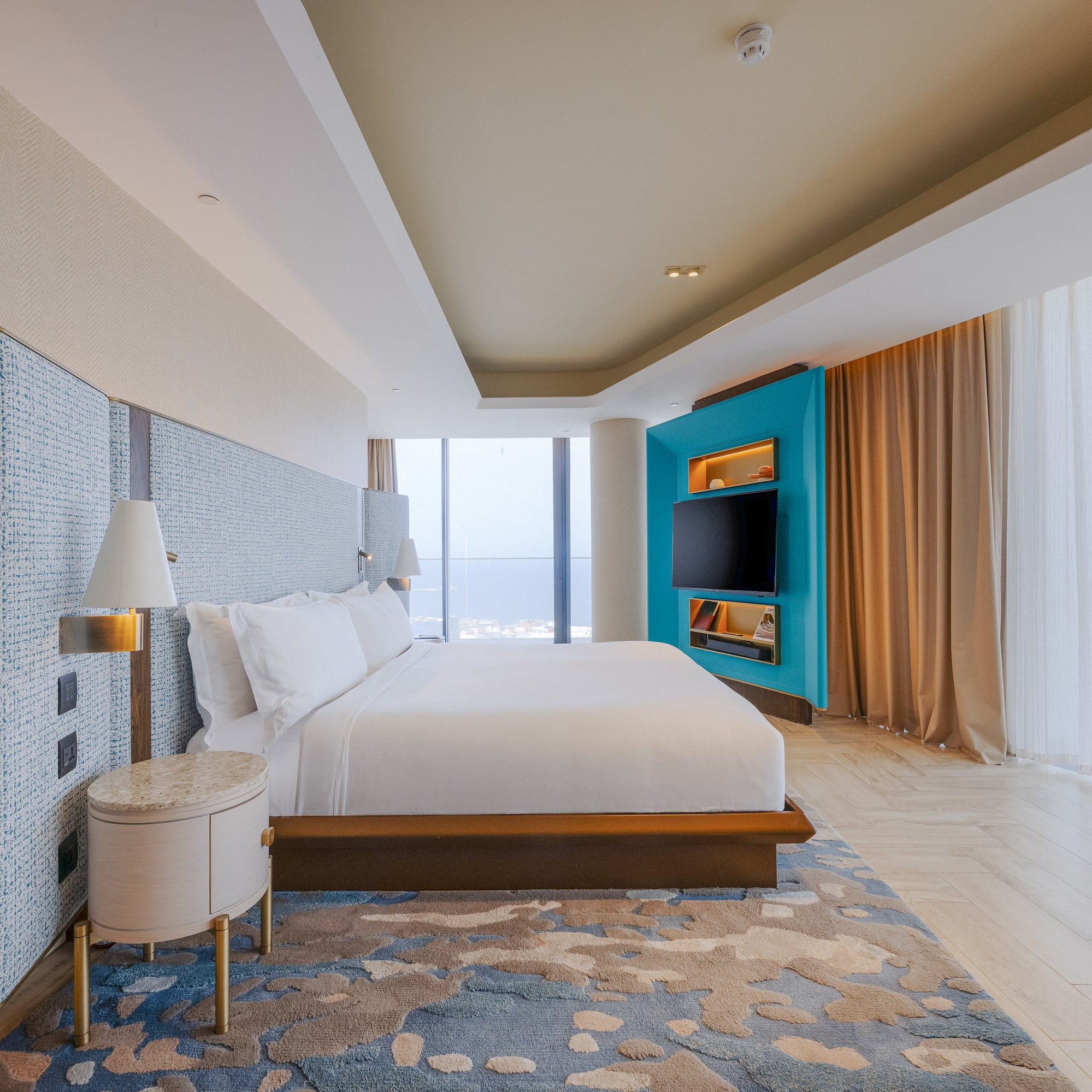 Luxury hotel bedroom with king bed, teal media wall, and panoramic city view.