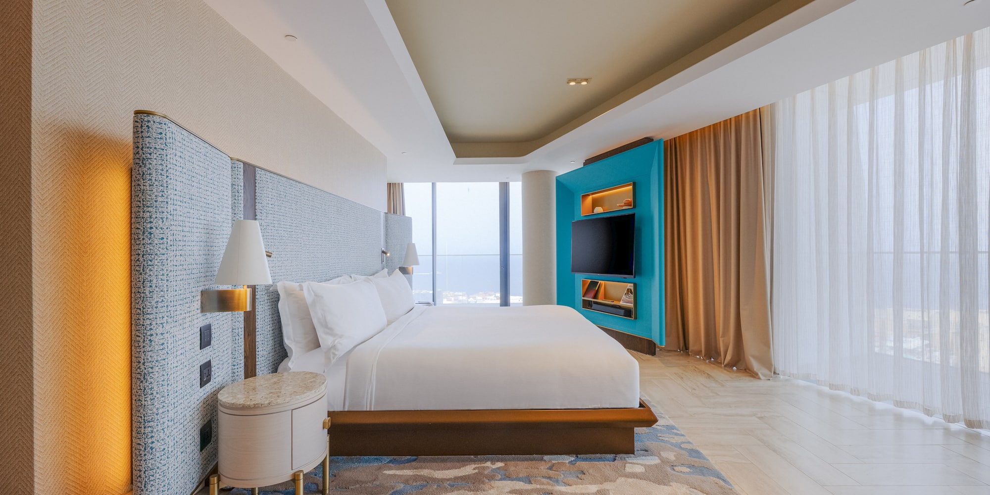 Luxury hotel bedroom with king bed, teal media wall, and panoramic city view.