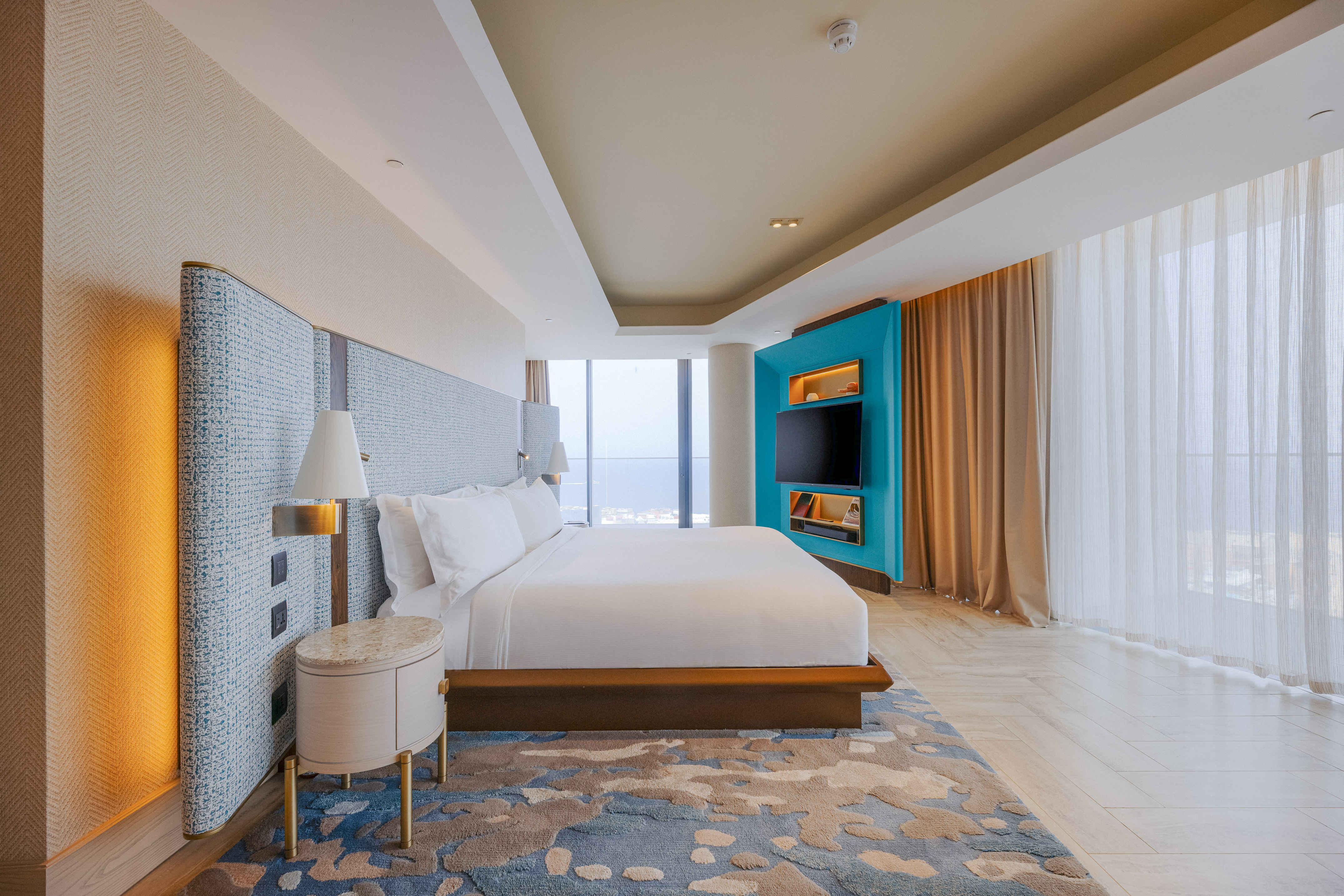 Luxury hotel bedroom with king bed, teal media wall, and panoramic city view.