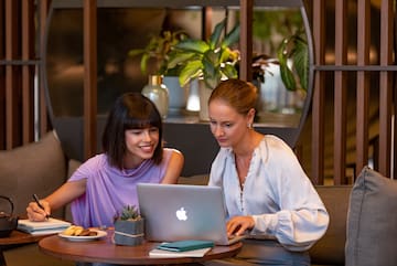 two women sitting at a table looking at a laptop