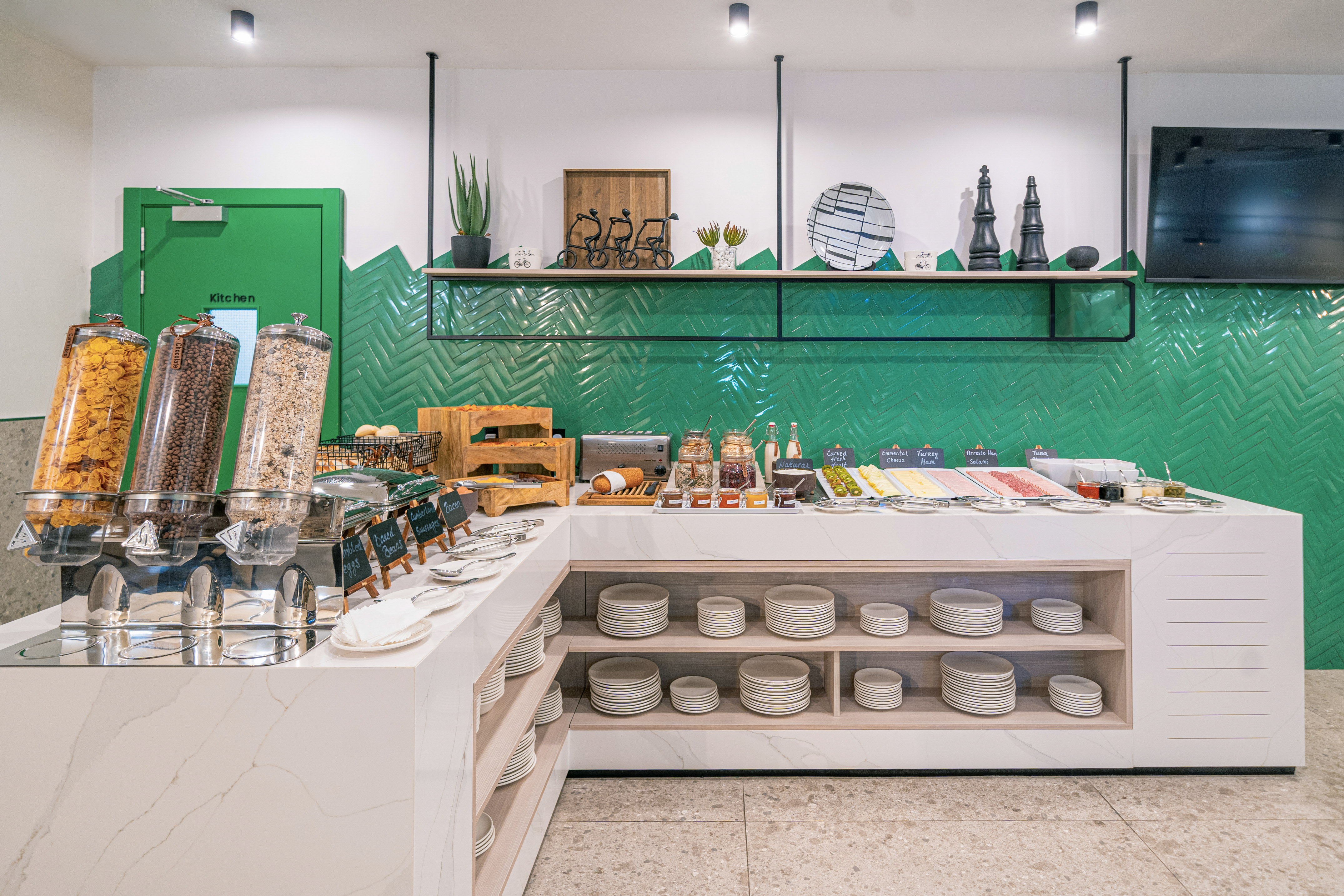 Modern breakfast buffet with cereals, hot food, deli, and green tiled wall.