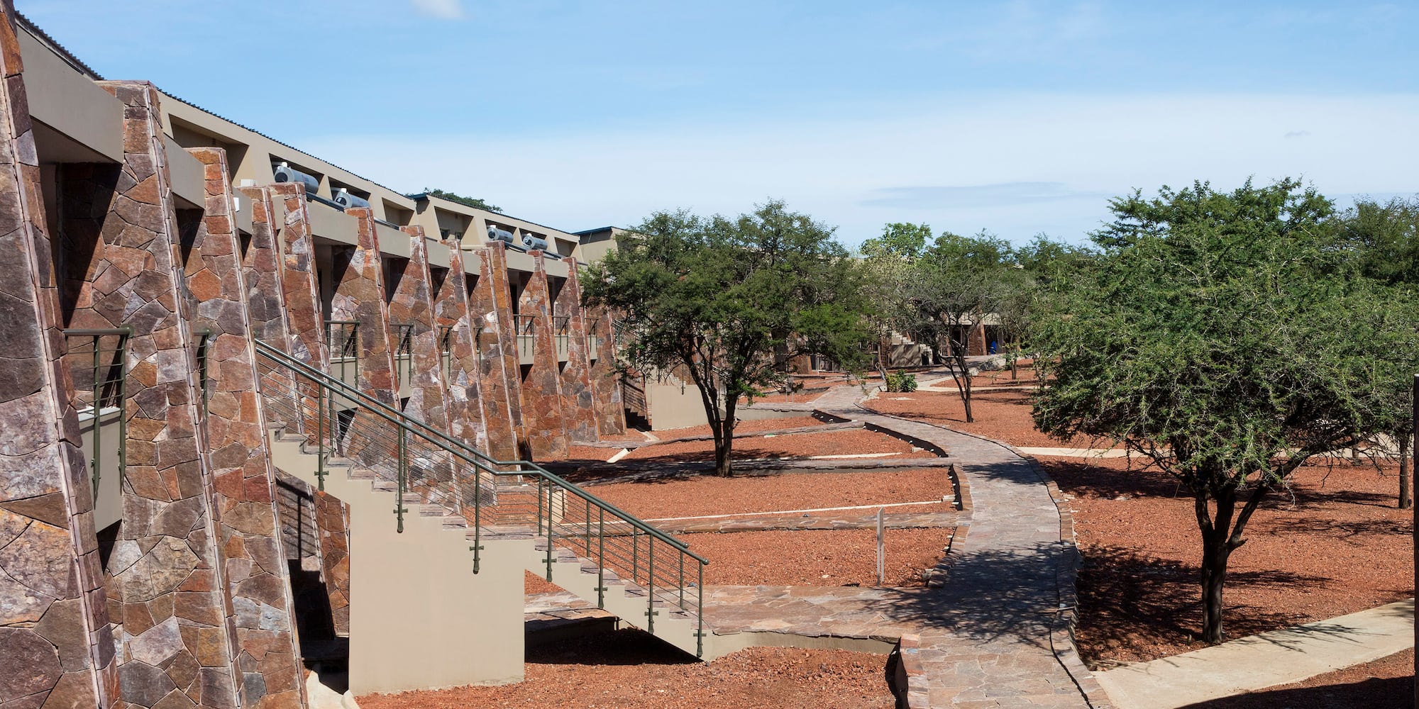 Stone-faced safari lodge, steps, winding paths, red earth, and green trees.