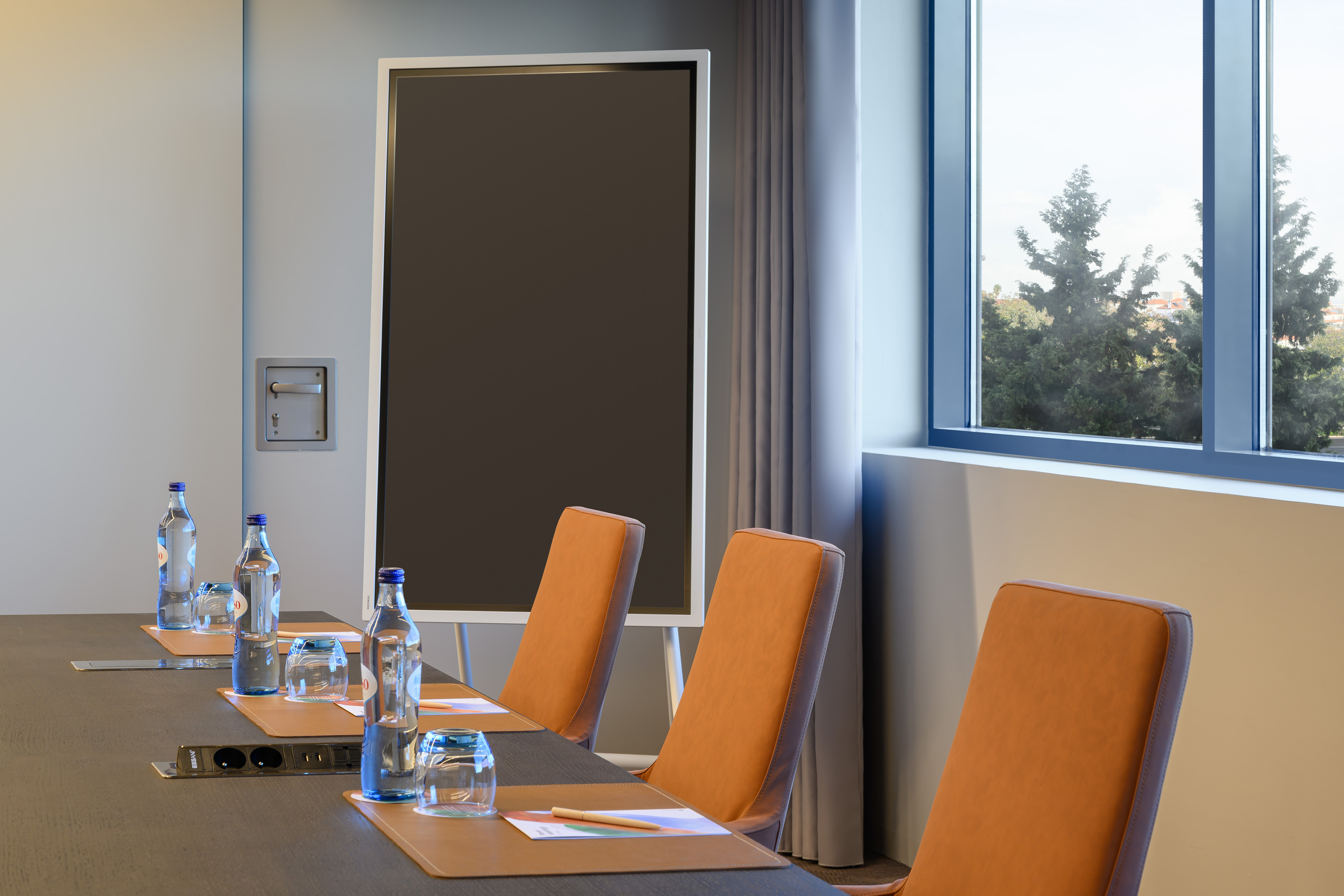 Modern conference room with dark table, orange chairs, water, and large screen.