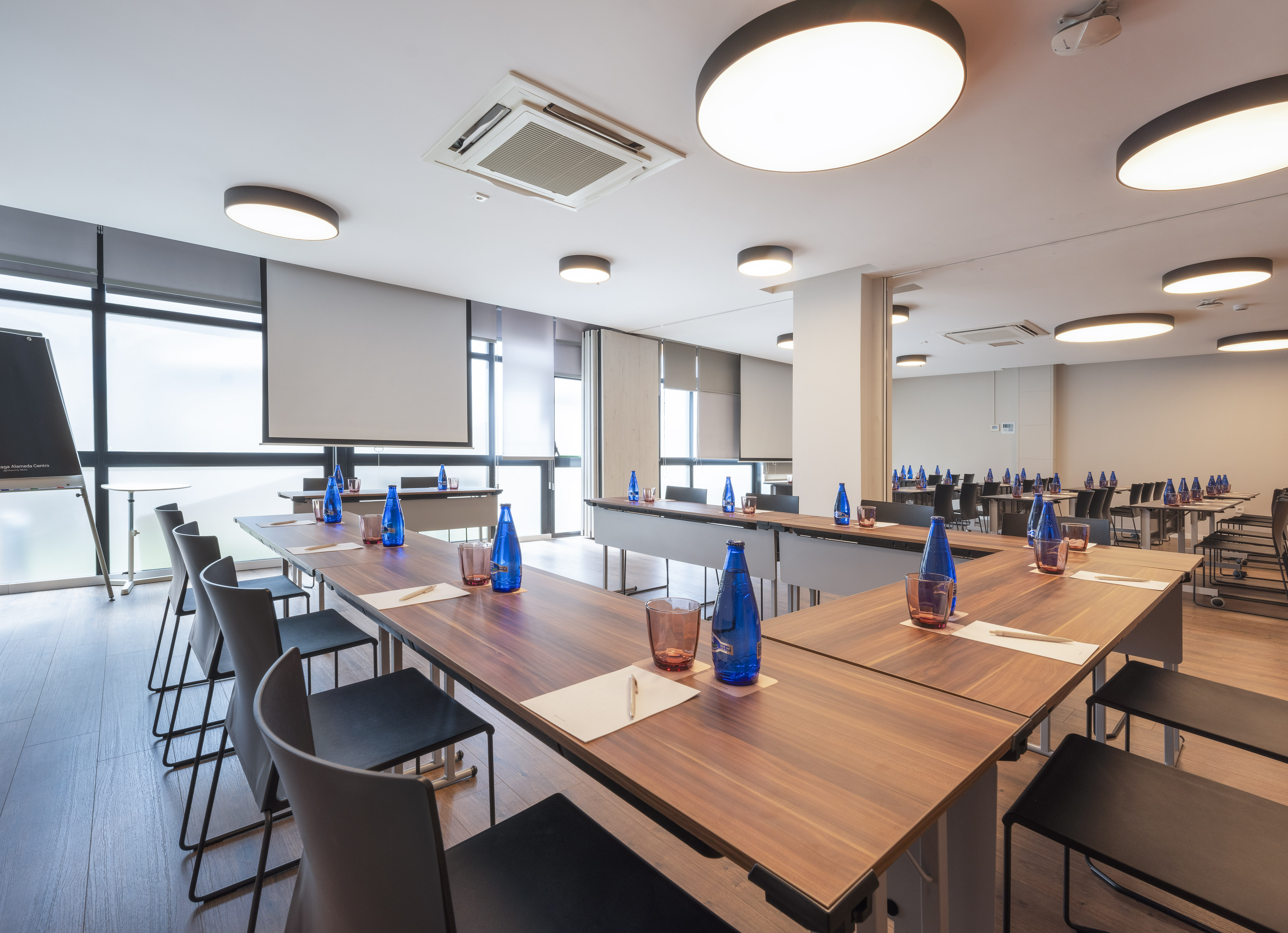 Modern conference room with U-shaped table, chairs, projector screens, and water bottles.