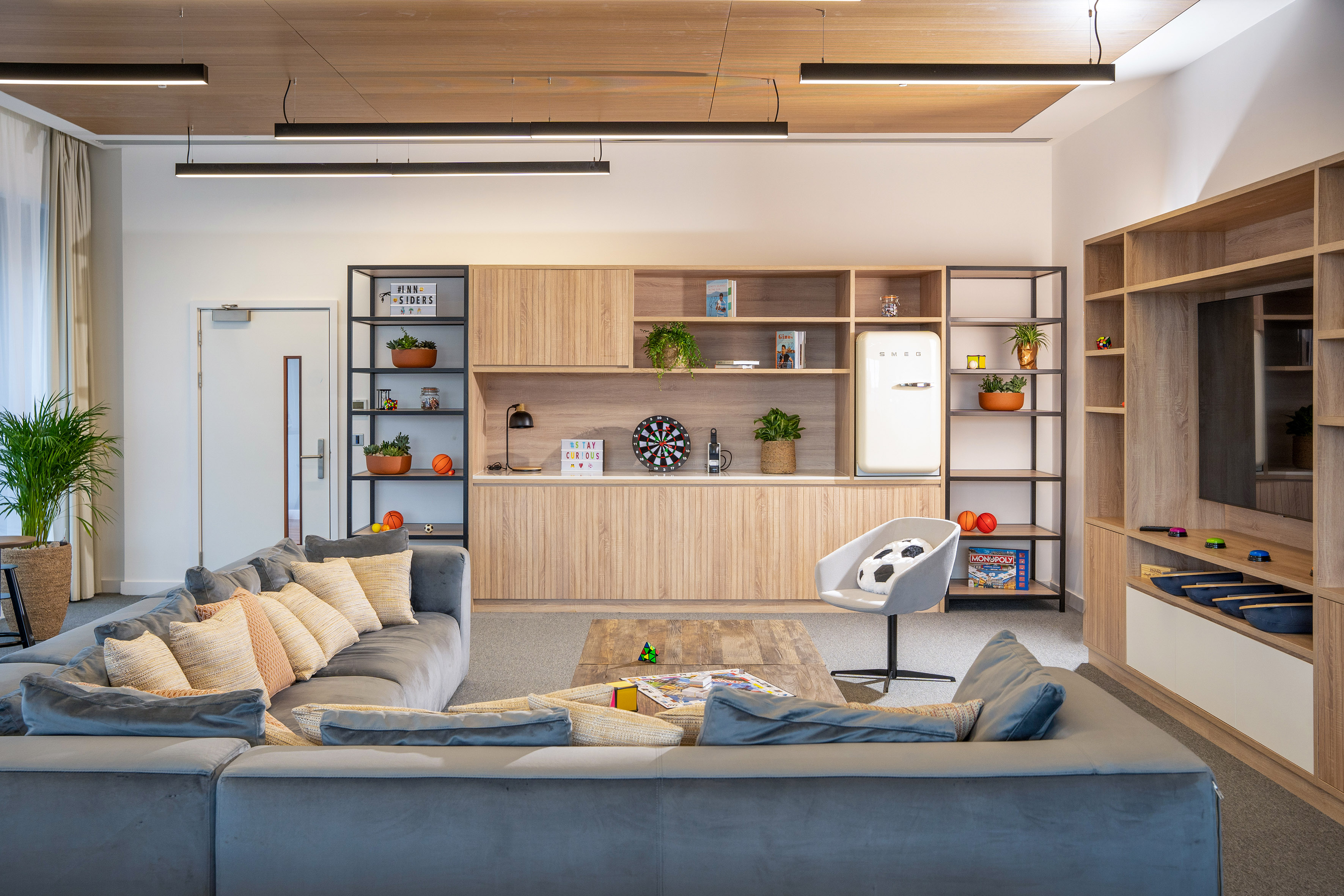 Modern lounge with sectional sofa, dartboard, Smeg fridge, and storage units.