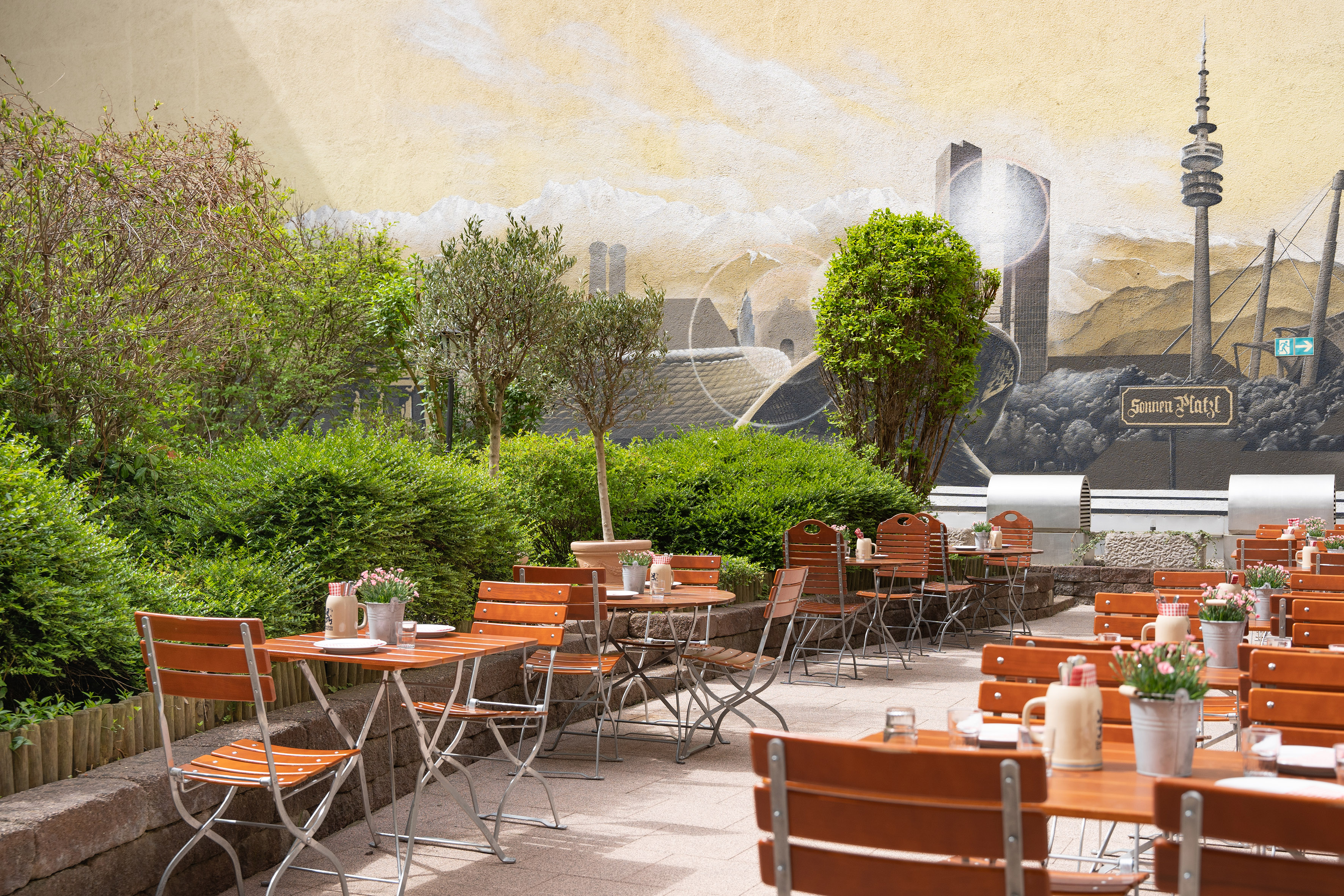 Beer garden patio with wooden tables, green plants, and city skyline mural.